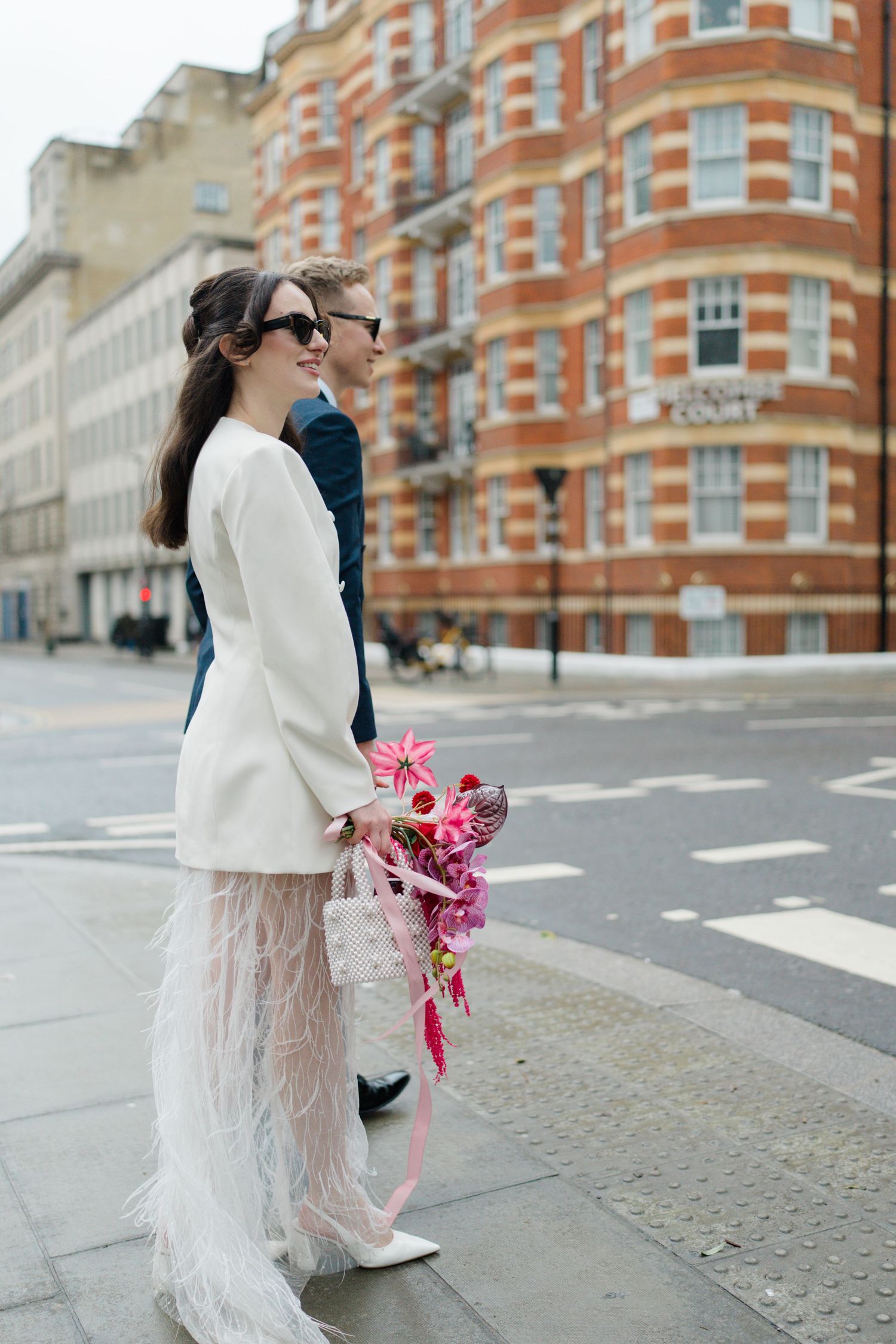 Bride in white blazer and feather skirt with pink bouquet and groom in navy suit crossing urban street in London.