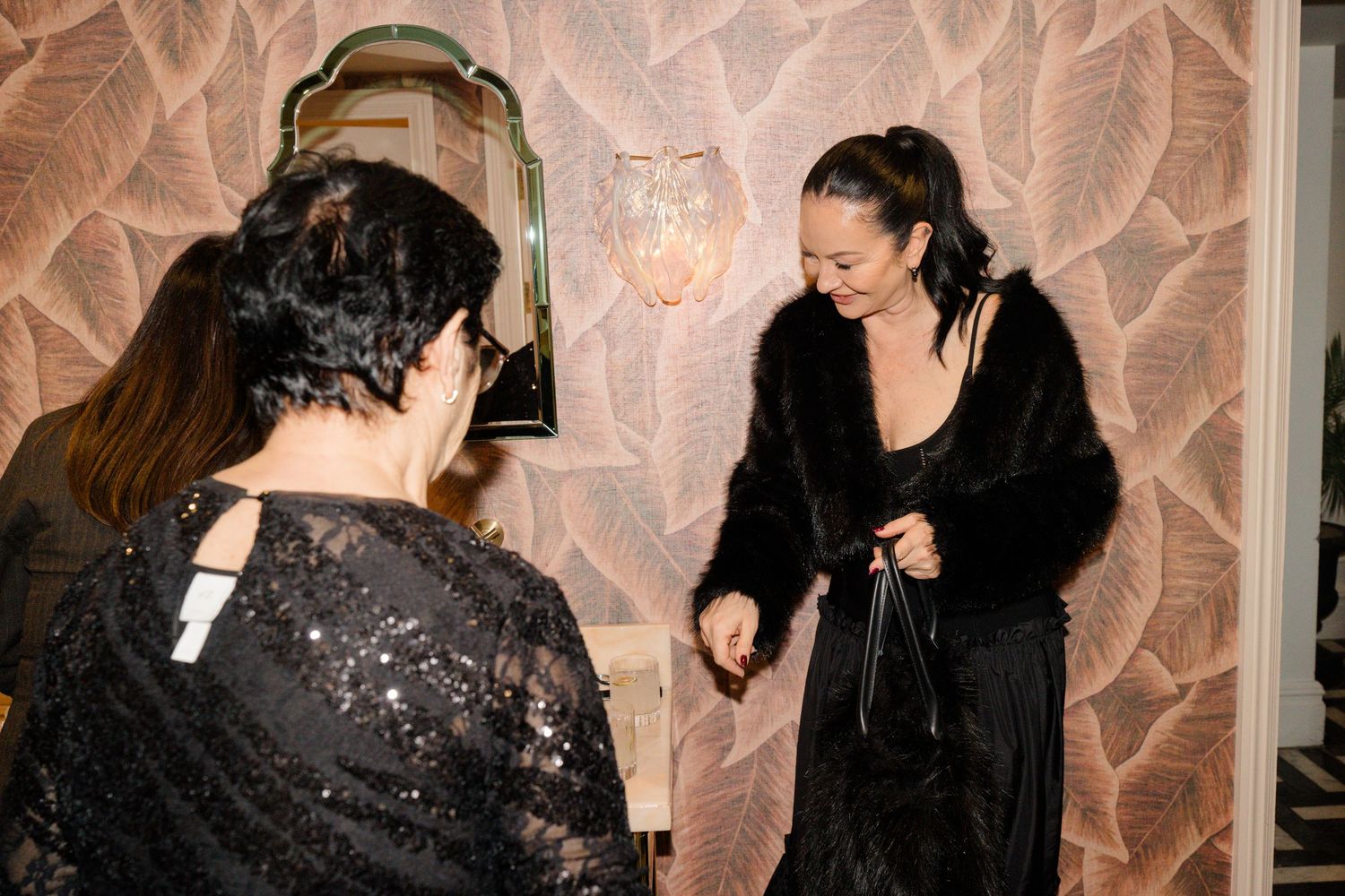 Woman in black fur coat adjusts outfit while guest in sequin dress looks on in floral wallpapered room.