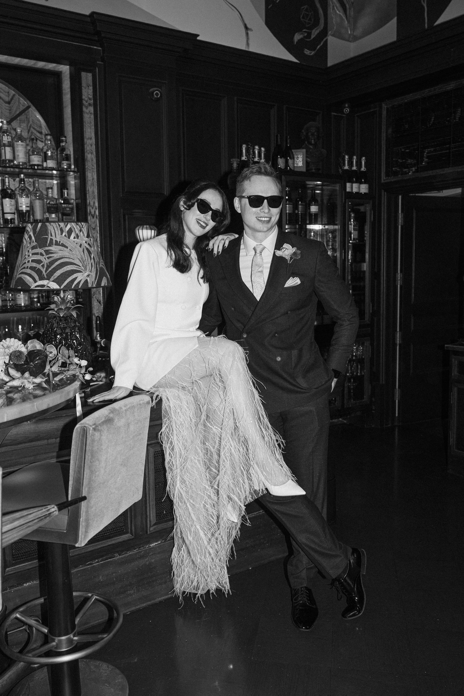 Black and white photo of a stylish bride and groom in sunglasses posing at a bar together.