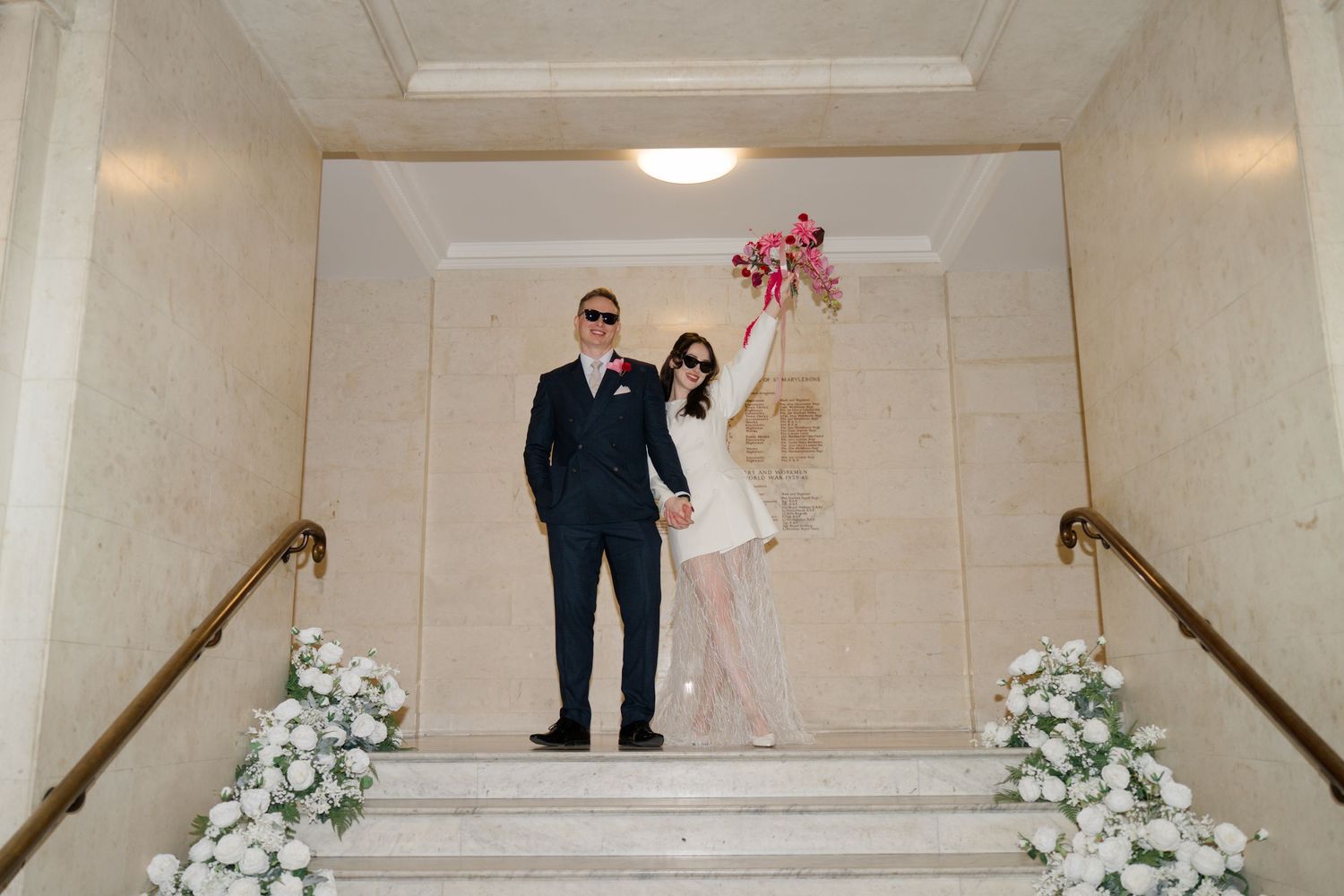 Bride and groom in sunglasses celebrate on marble staircase decorated with white flowers, bouquet raised.