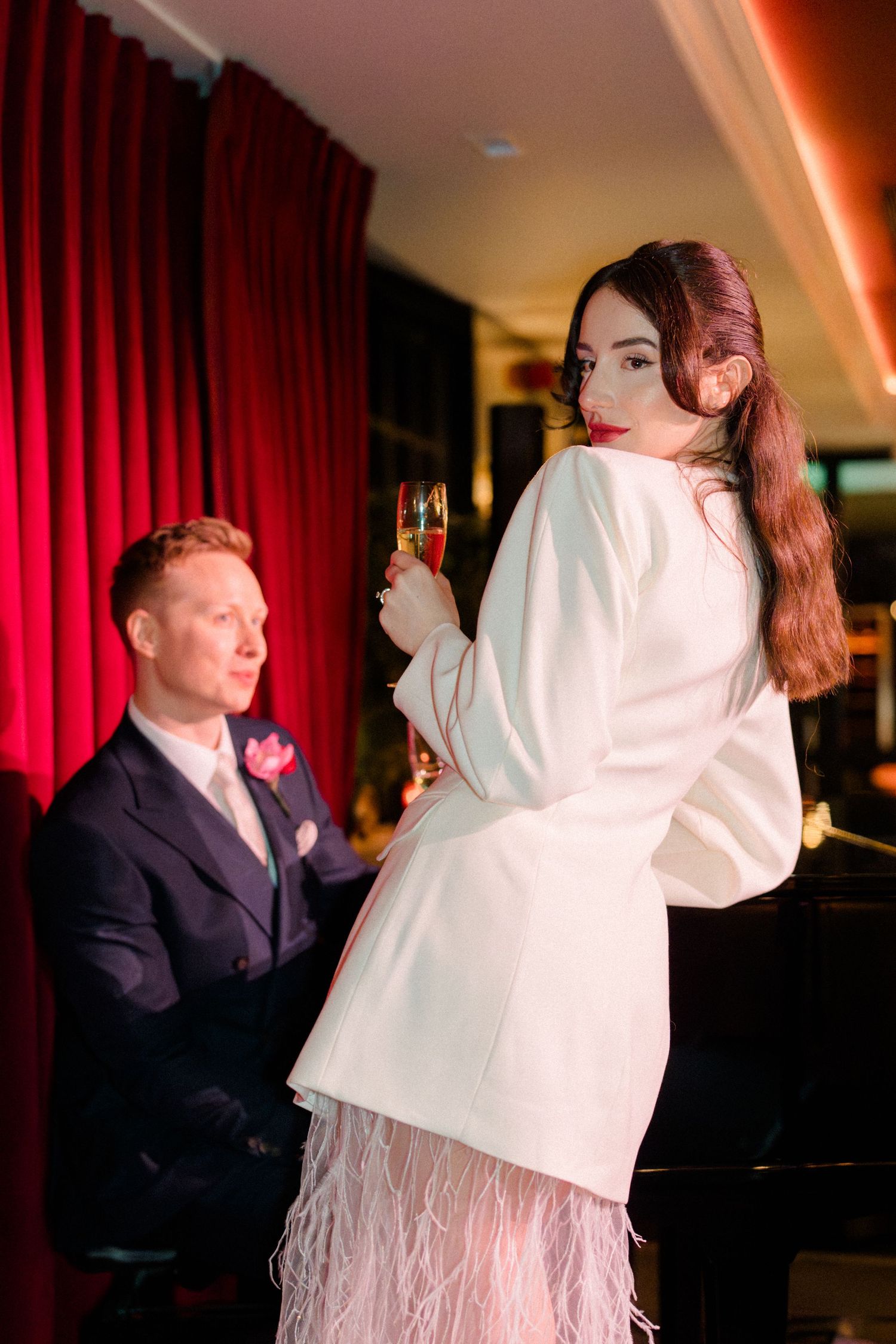 Bride in white feathered mini dress holds champagne glass beside groom at piano bar with red curtains.