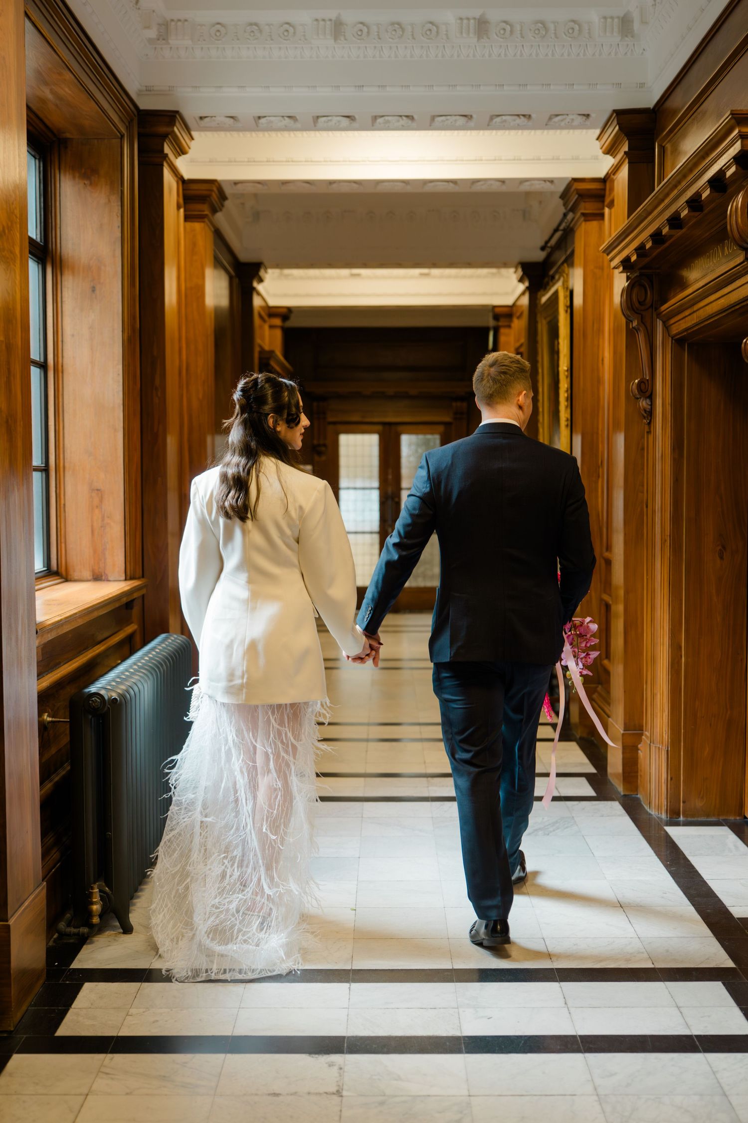 Bride and groom walking hand in hand through elegant wood-paneled hallway with marble floor.