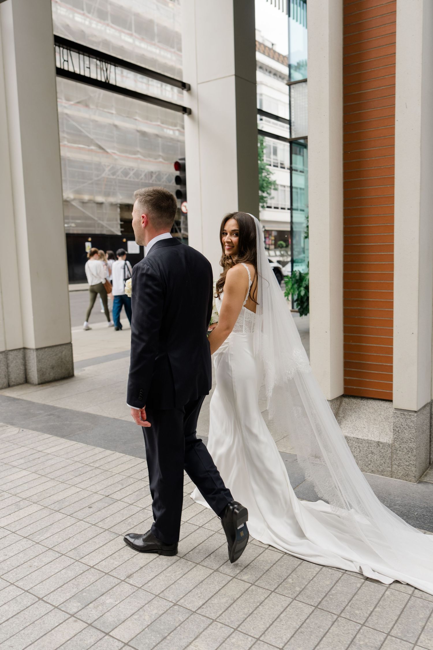 Bride in flowing white gown and veil walks with groom through city streets on wedding day.