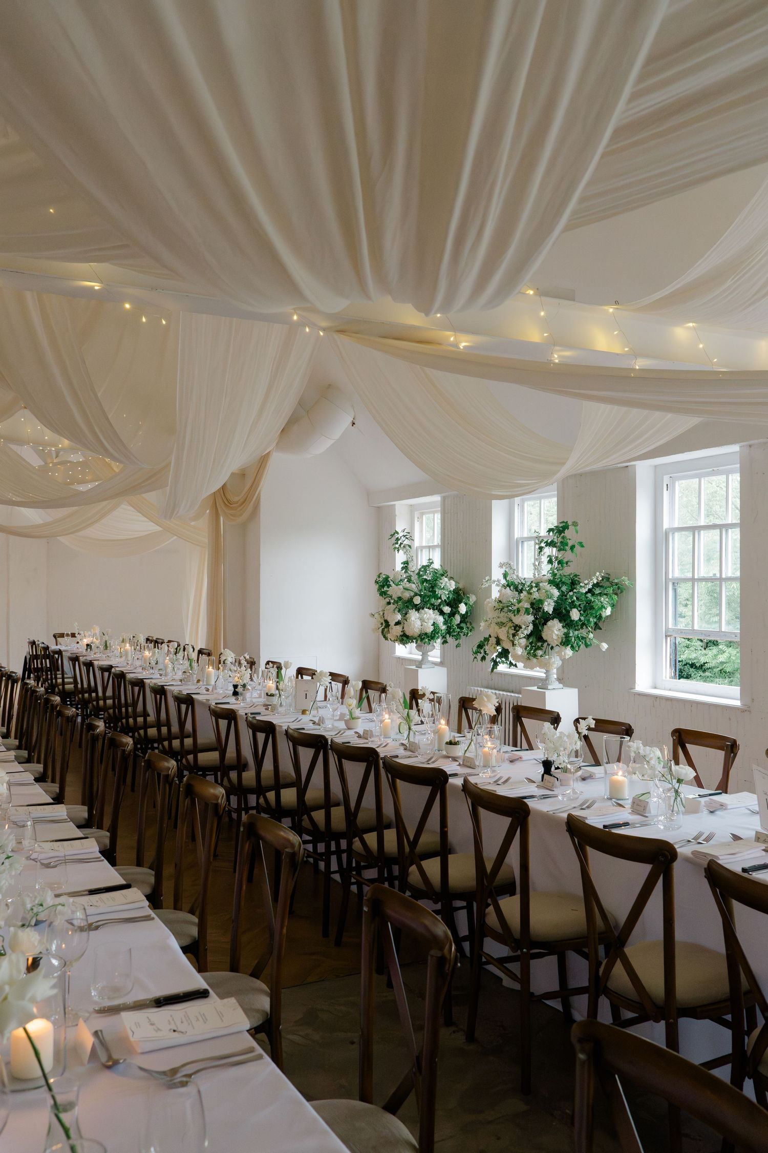 Elegant wedding reception hall with white draped ceiling, fairy lights, long tables, dark cross-back chairs, and tall floral centerpieces.