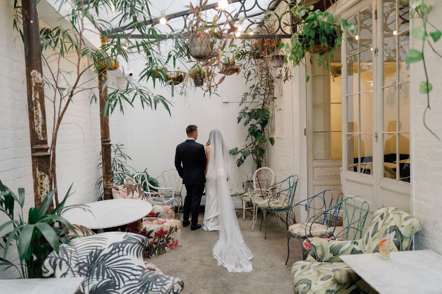 Bride and groom share a private moment in a lush botanical courtyard adorned with hanging plants and greenery.