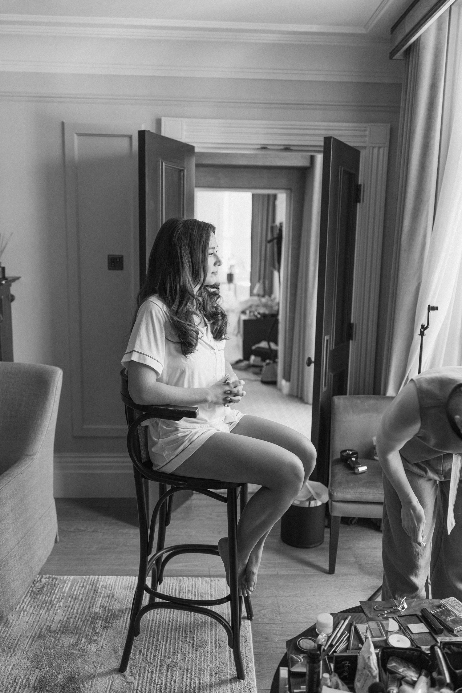 Black and white photo of bride in white robe sitting on chair before mirror during wedding prep.