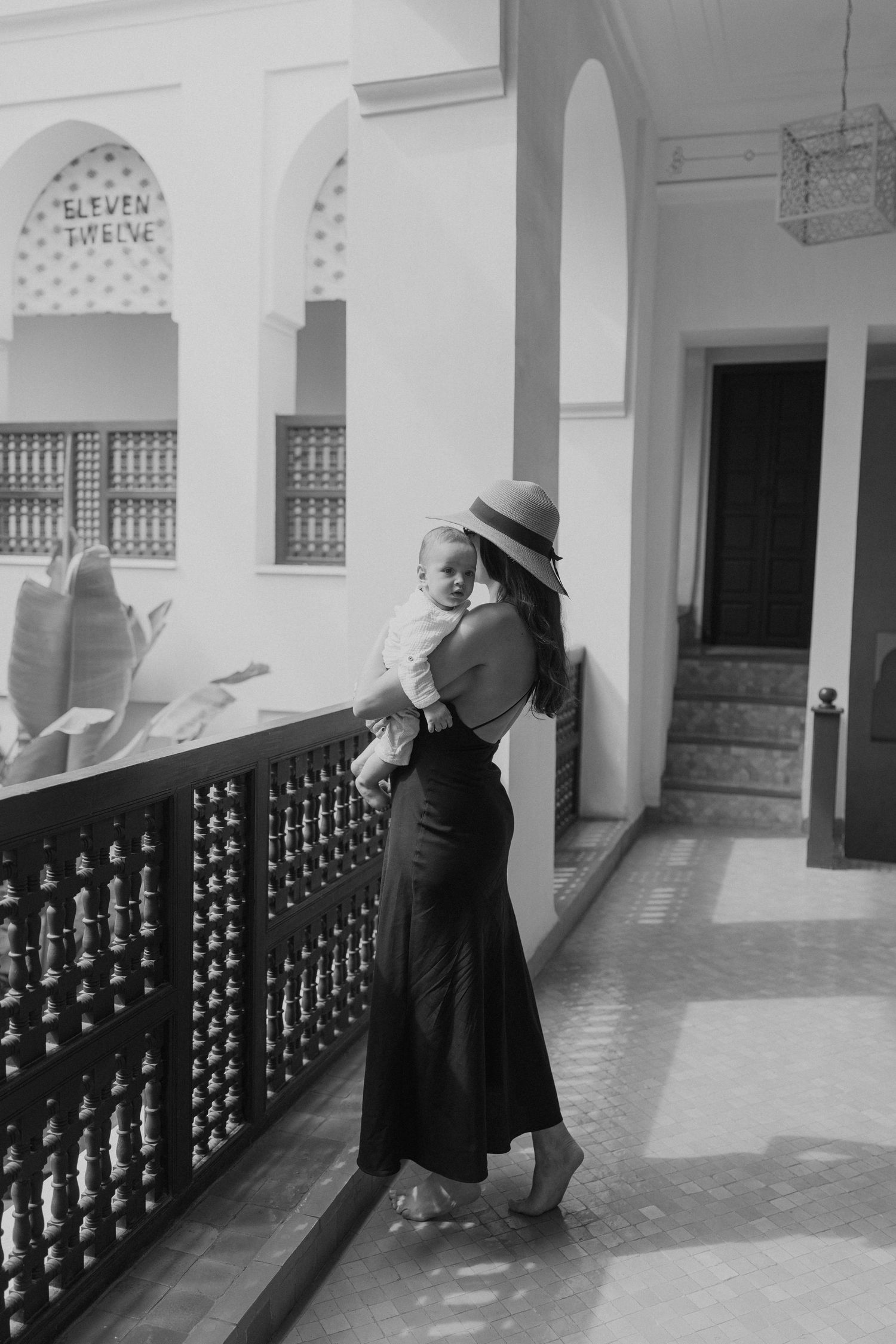 Black and white photo of a woman in a hat holding a baby on a balcony of a Moroccan riad.
