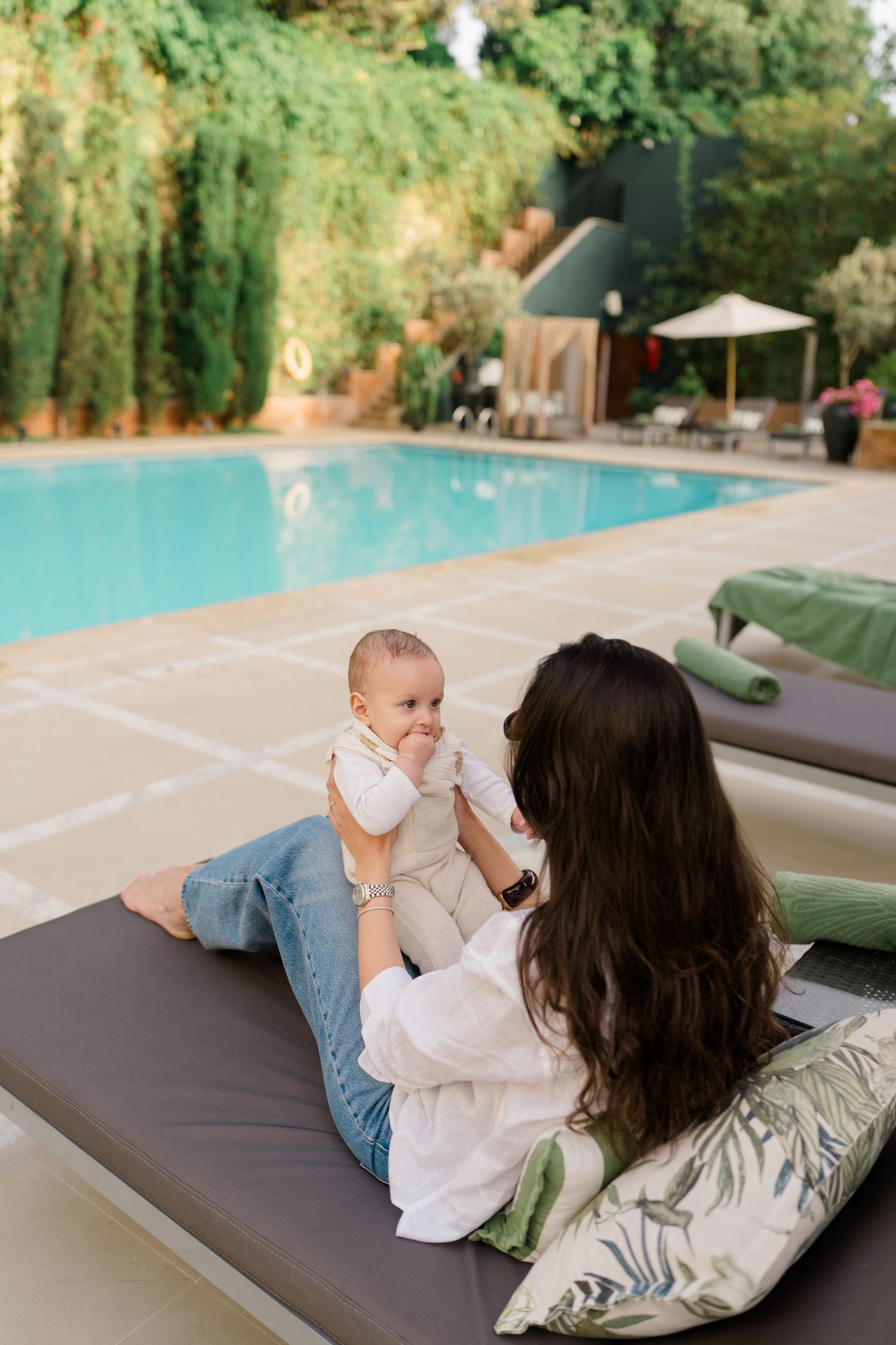 Mother holding smiling baby on poolside lounge chair, lush greenery and luxury resort pool in background.