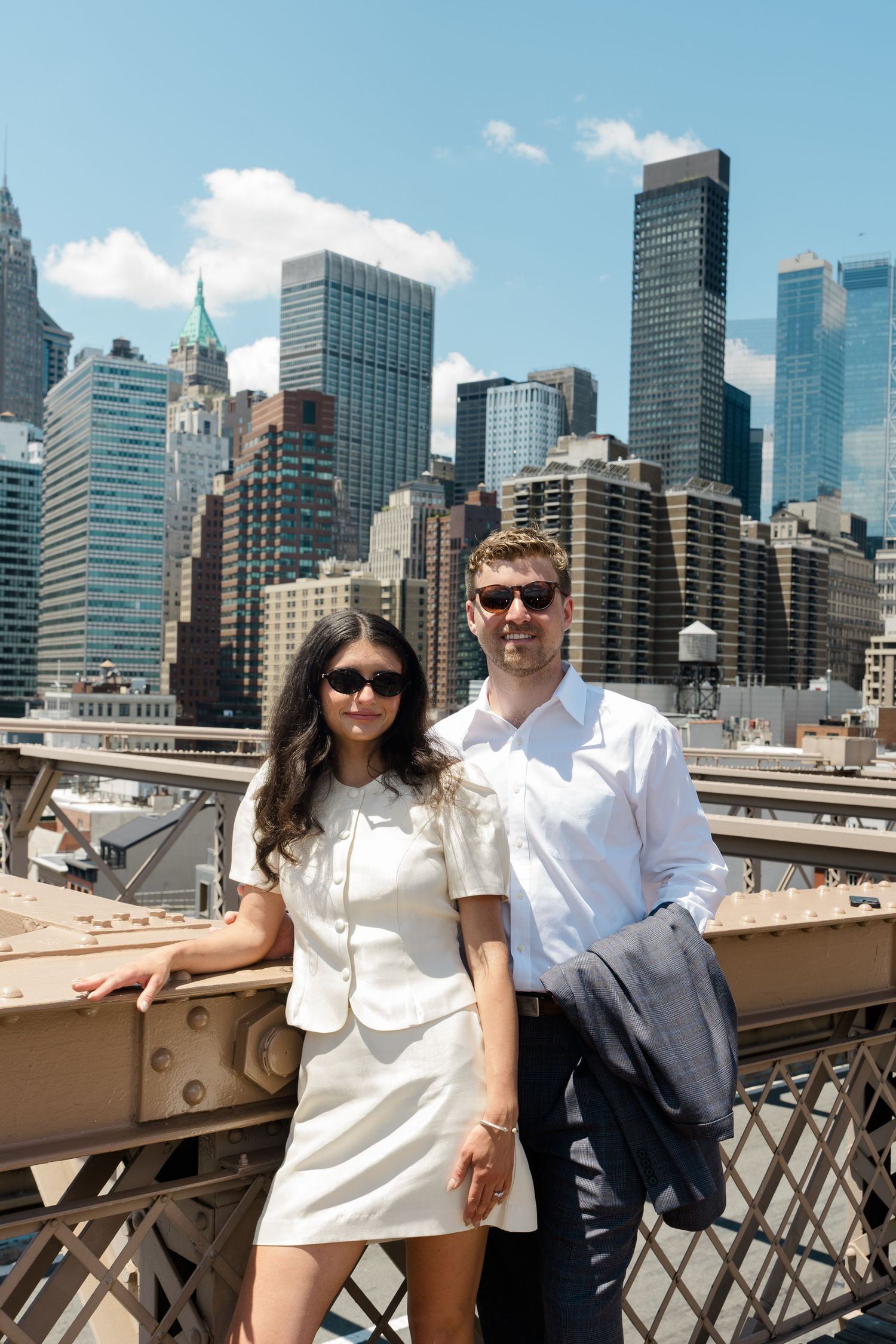 Couple in white outfits and sunglasses posing on Brooklyn Bridge with Manhattan skyline in background on sunny day.