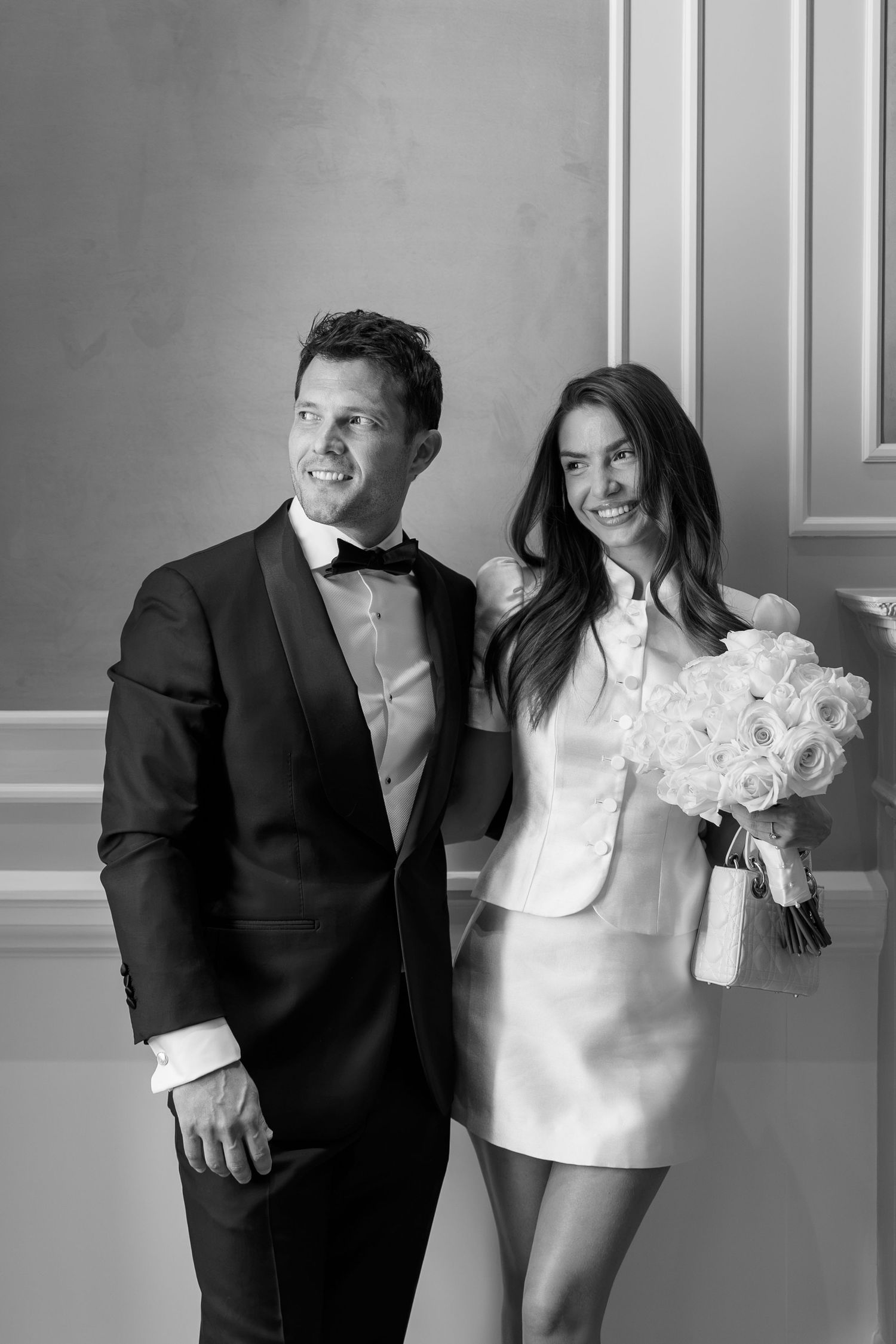 Black and white photo of a smiling couple in formal attire, groom in tuxedo, bride in mini dress holding white roses.
