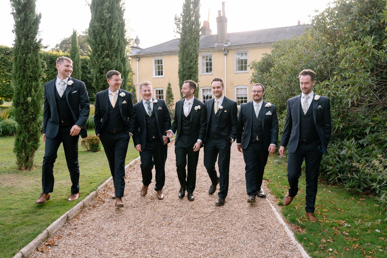 Wedding party in matching suits walking together on a gravel path surrounded by cypress trees.