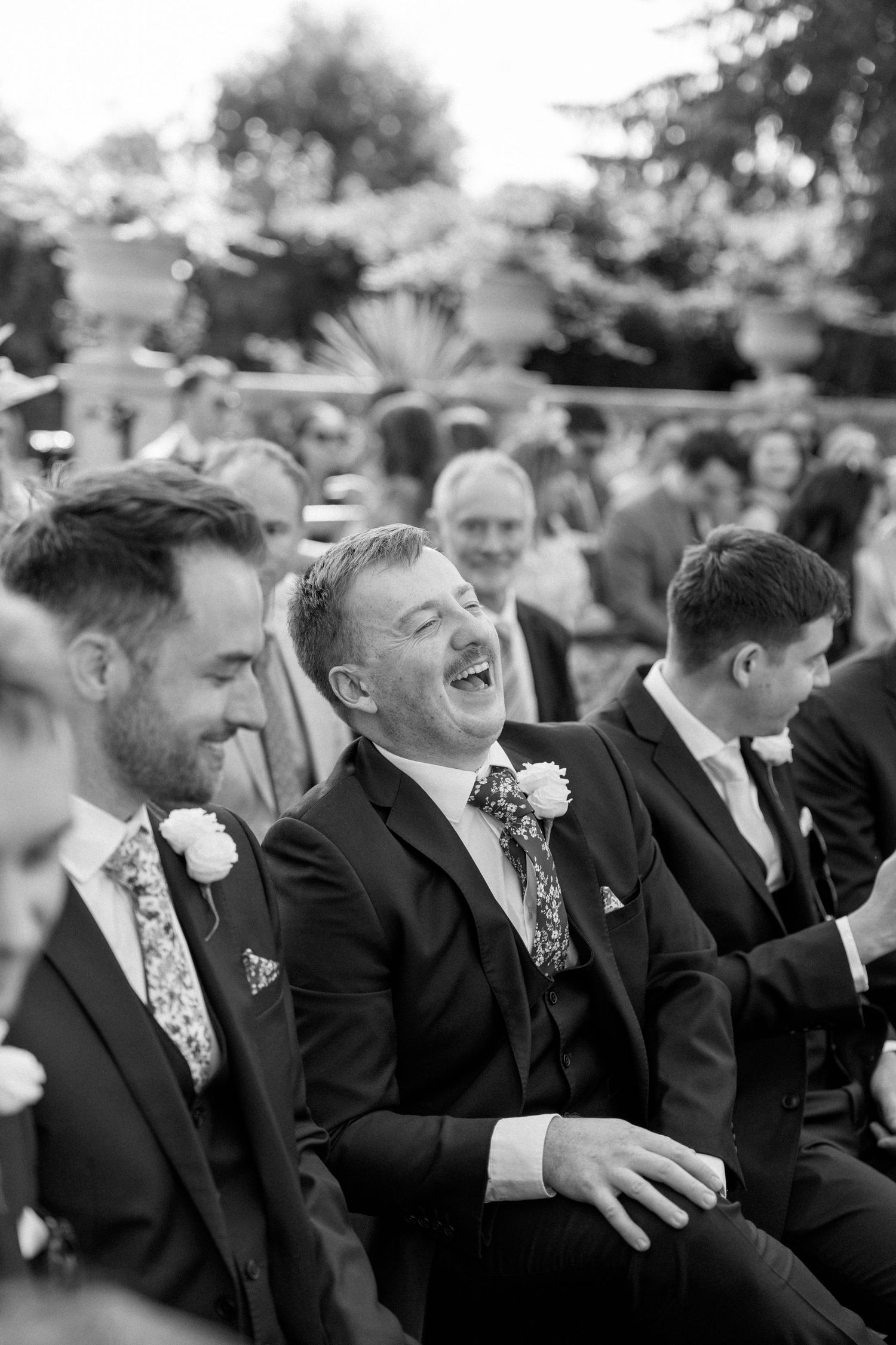 Wedding guests in formal attire laugh joyfully during outdoor ceremony.