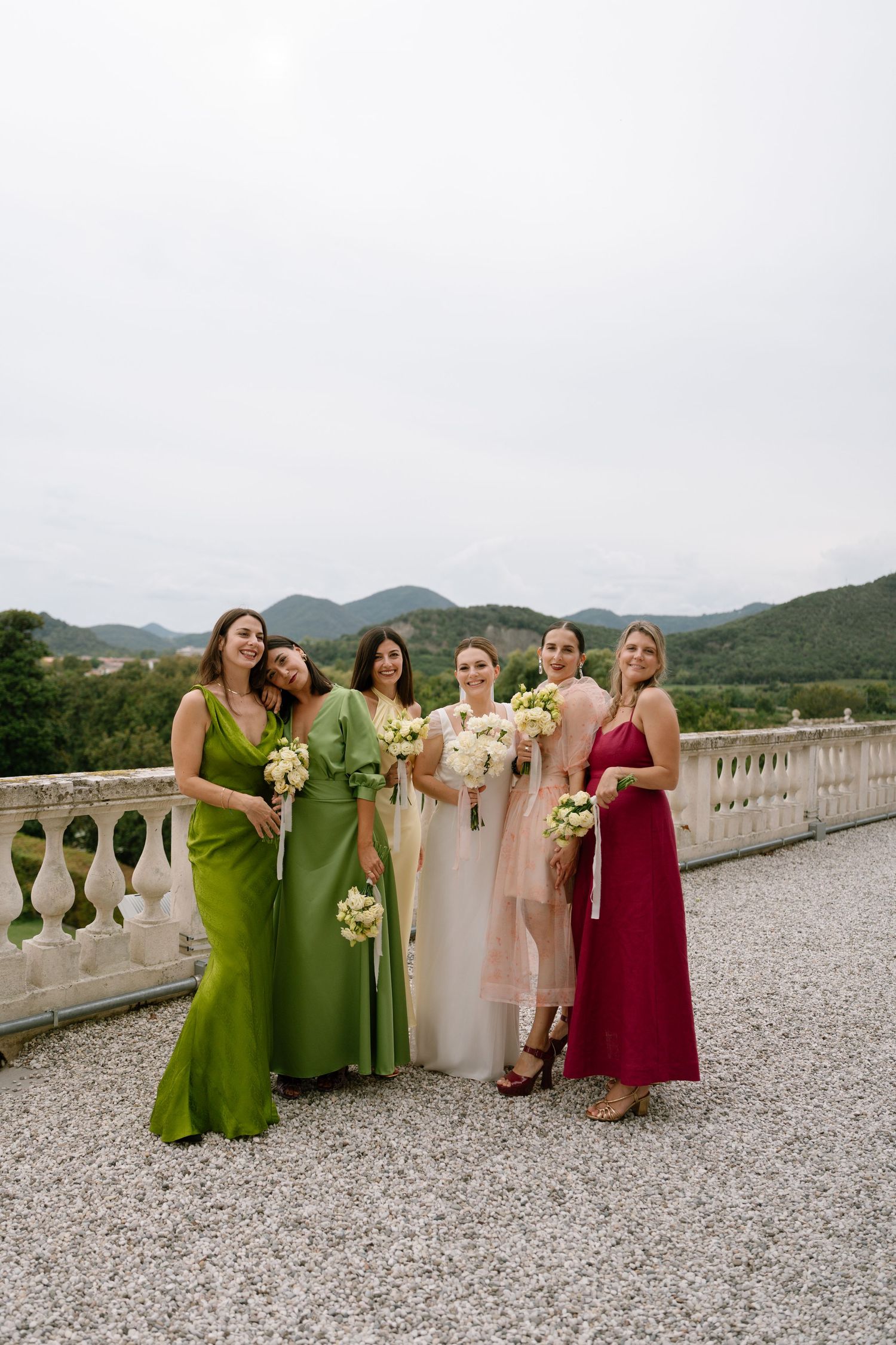 Bridesmaids in coordinating green, pink and burgundy dresses gather for photos on a terrace.