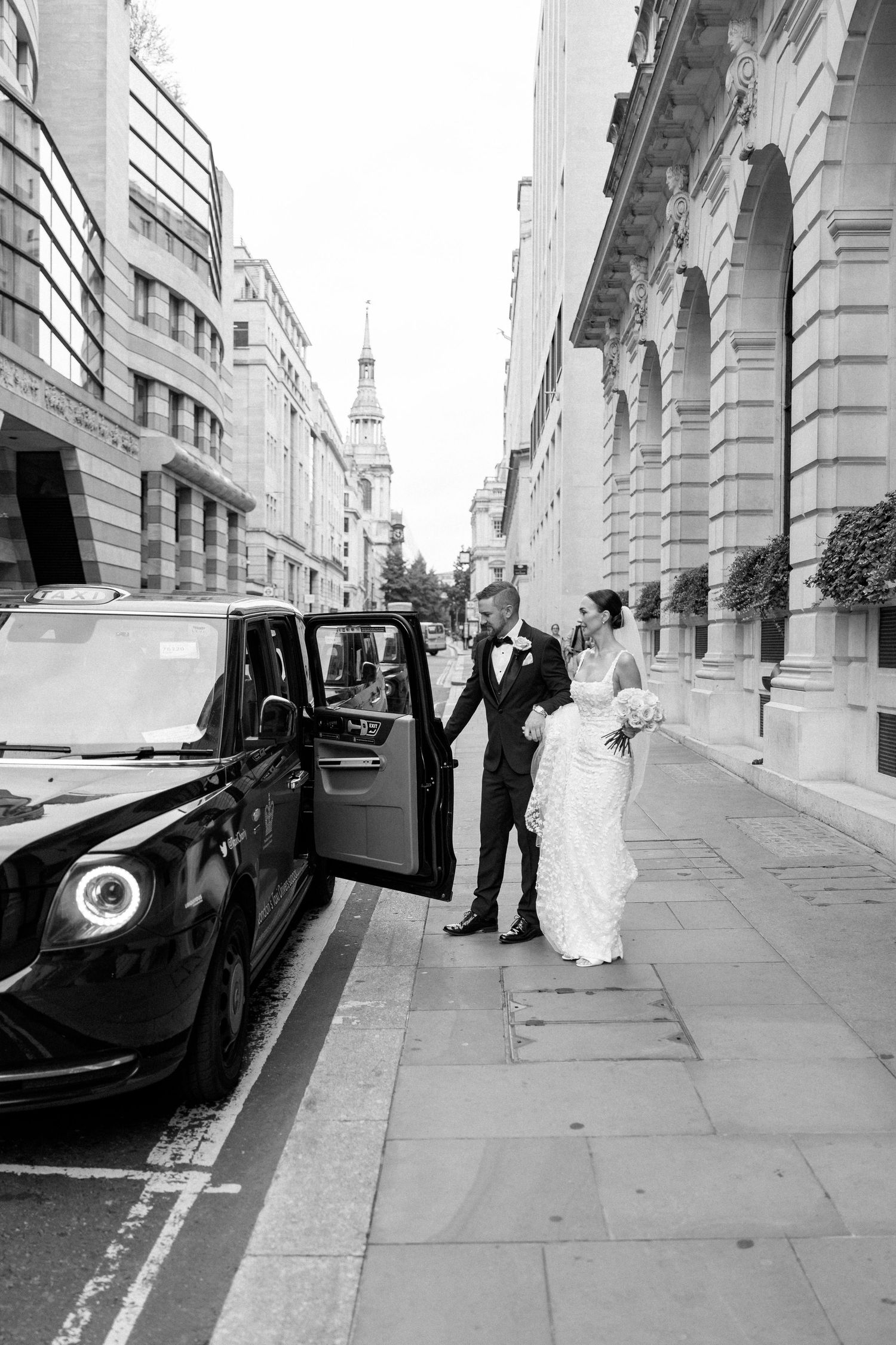 Black and white photo of a couple next to a luxury car with London architecture in the background.