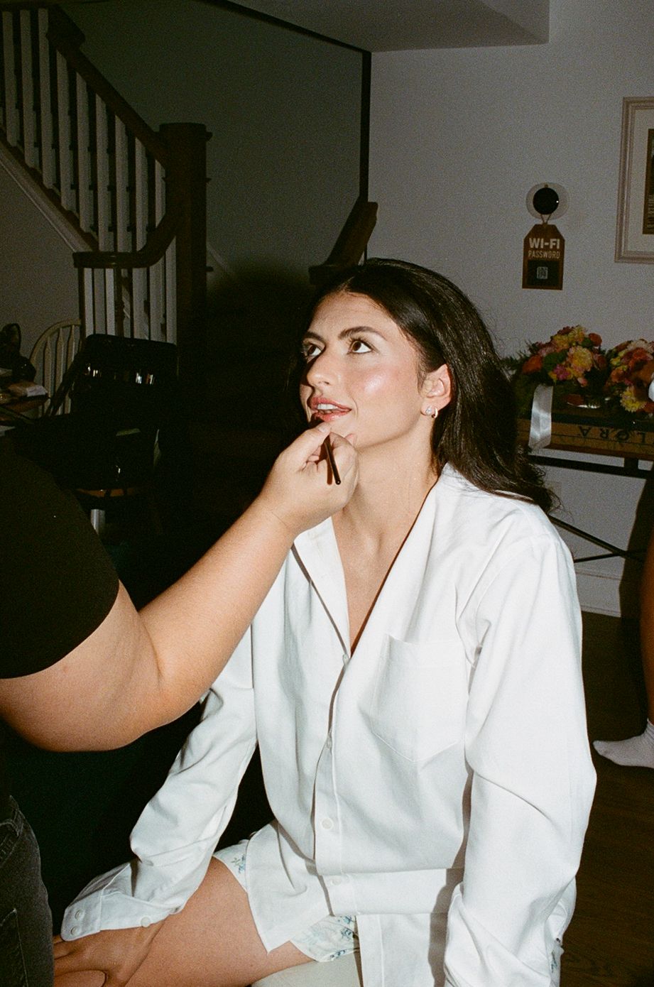 A young woman in a white robe sits getting makeup applied, looking up thoughtfully in a warmly lit home.