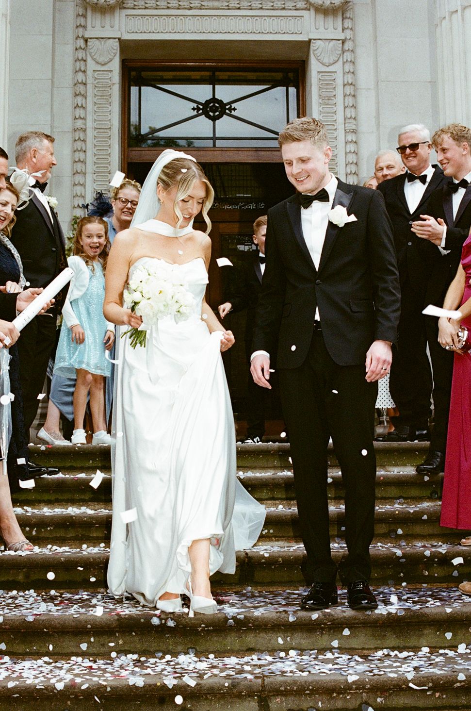Bride and groom exit church steps in confetti shower, she in white gown with veil, he in black tuxedo.