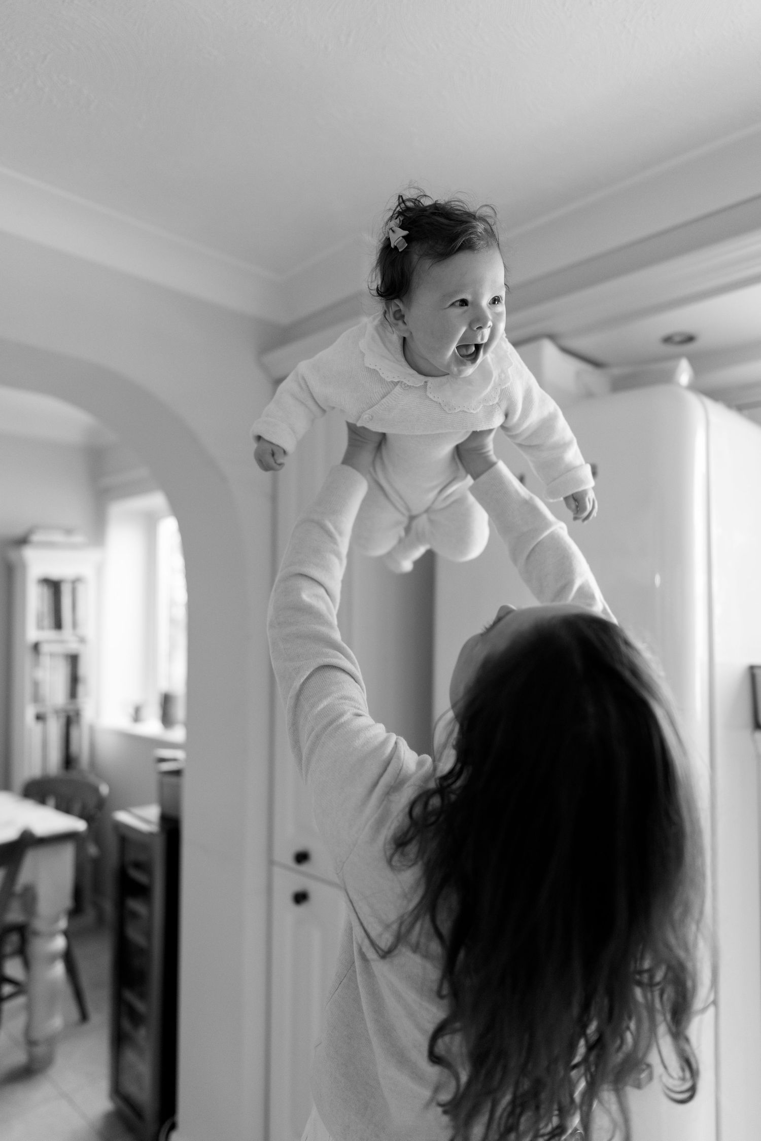 Mother lifting happy baby in white outfit against bright interior.