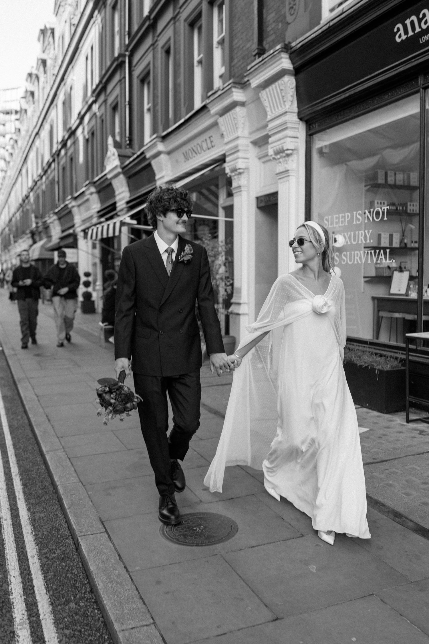 Wedding couple walking along a city sidewalk past various retail storefronts.