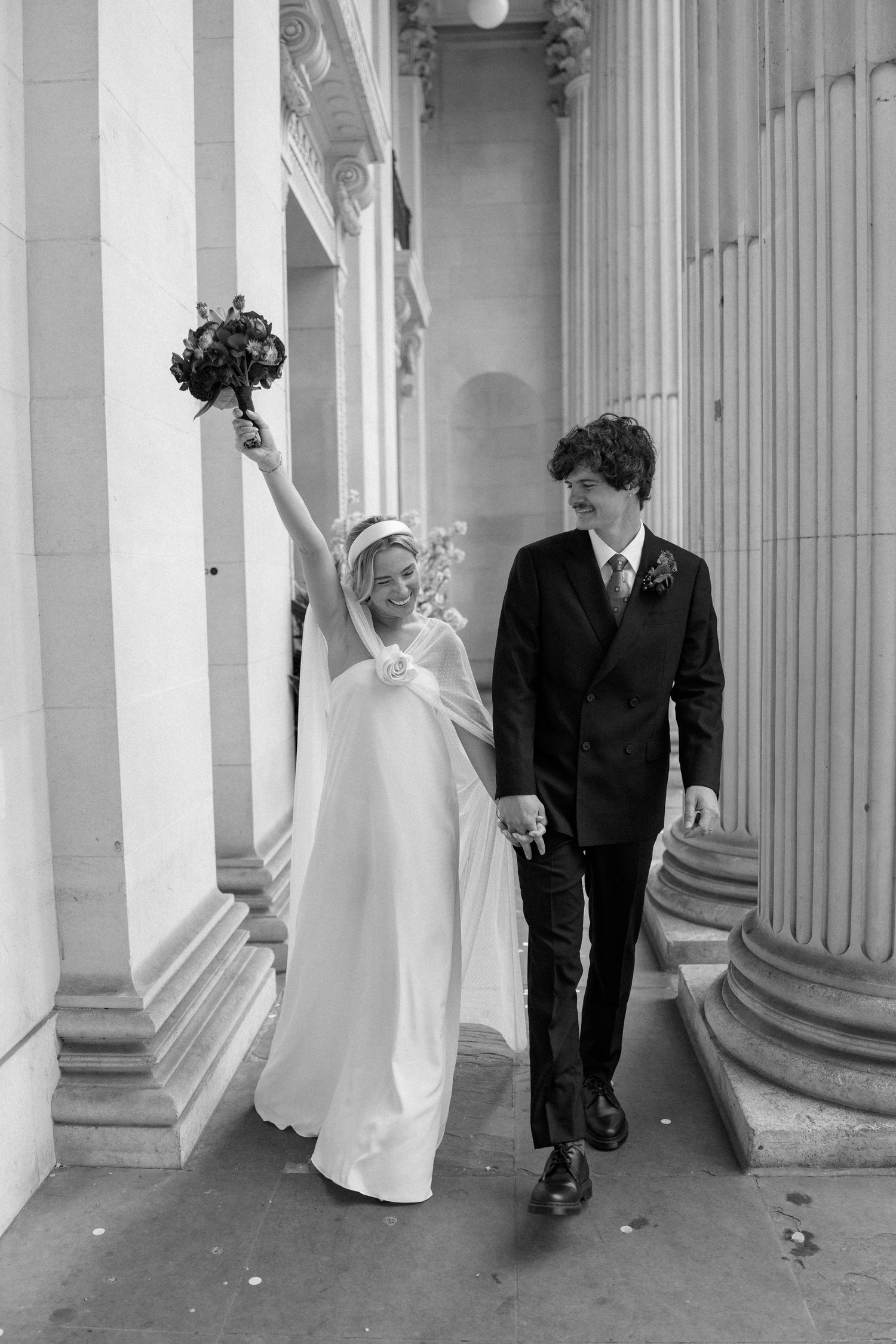 A joyful wedding couple walking between classical columns with the bride raising her bouquet.