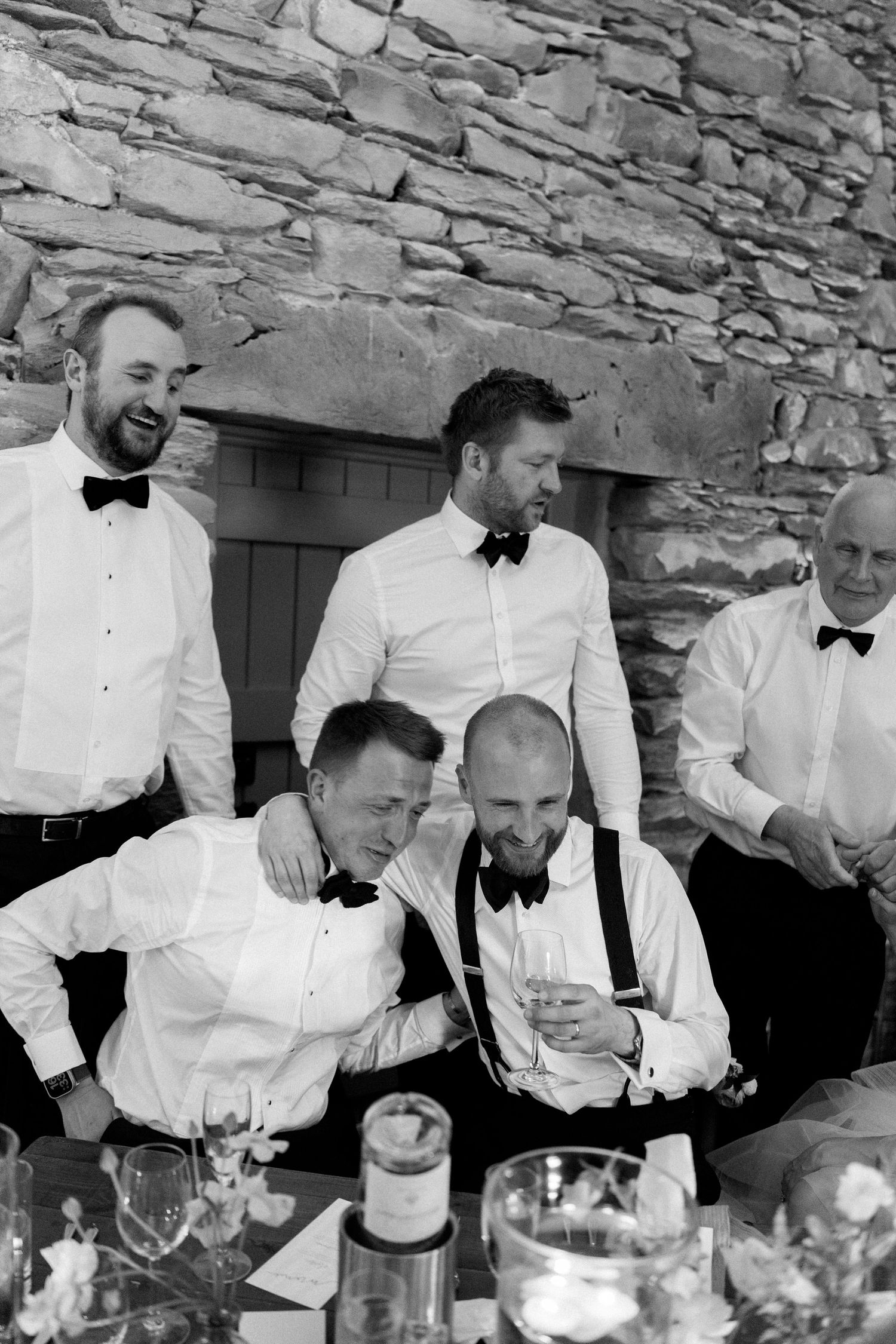 Groomsmen in formal attire sharing drinks and conversation at a stone-walled venue.