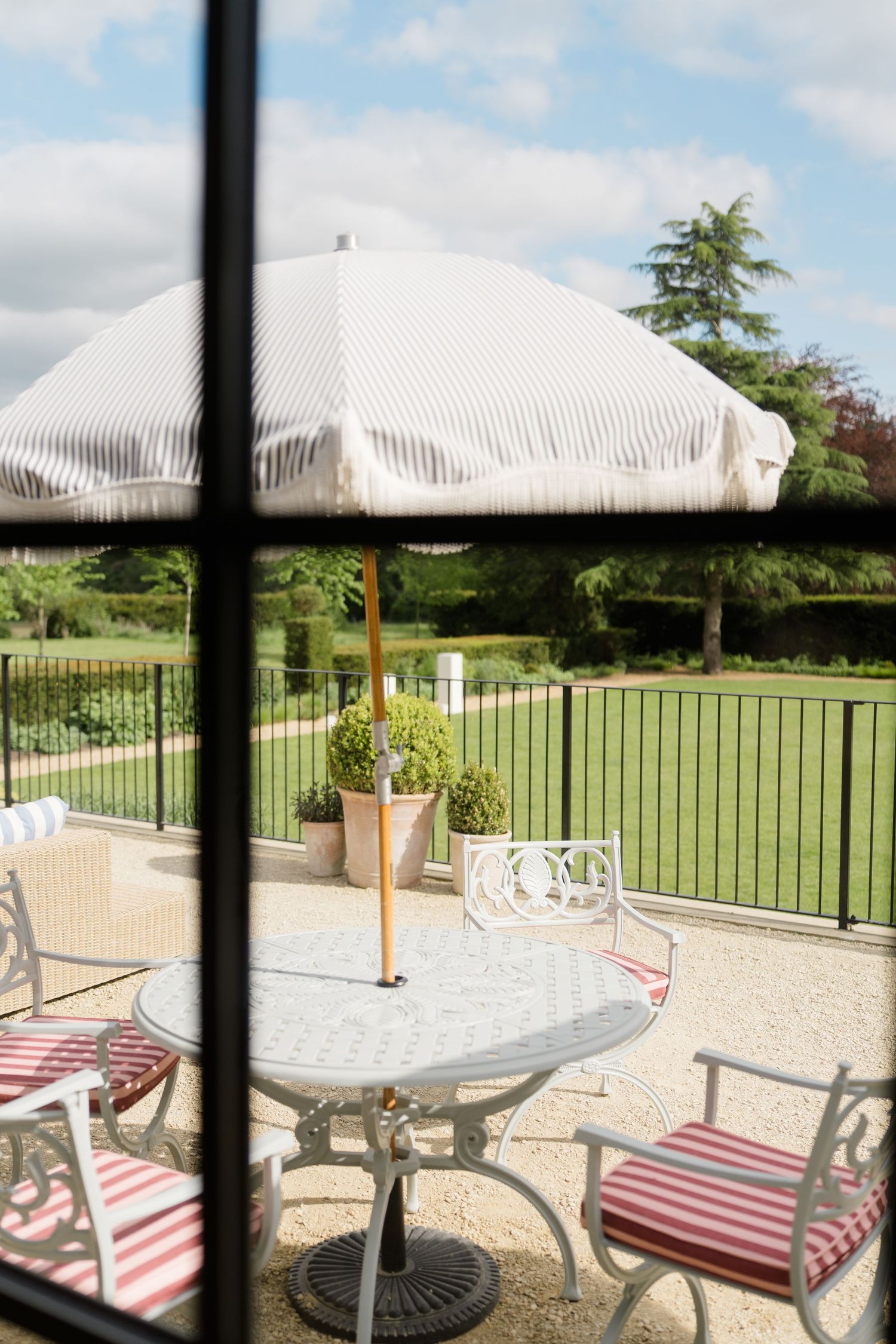 Elegant outdoor patio with white striped umbrella, iron furniture and manicured garden views.