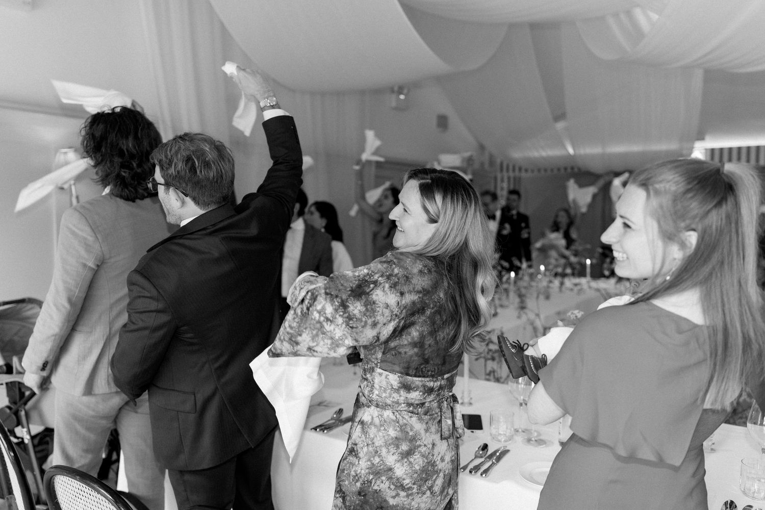 Black and white photo of wedding guests dancing and celebrating joyfully inside a draped reception marquee.