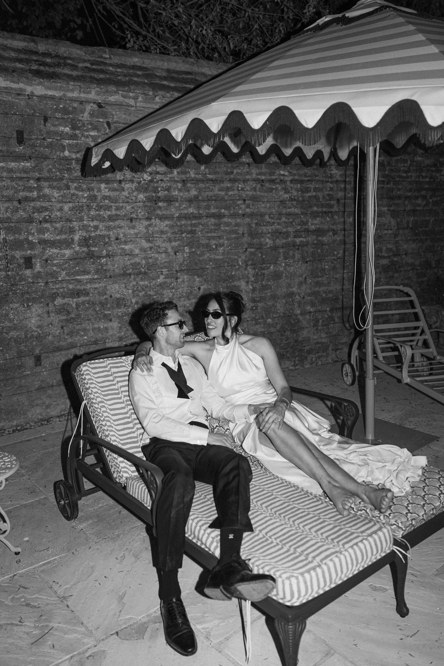 Bride and groom relax on striped chaise lounge under canopy in black and white evening photo.