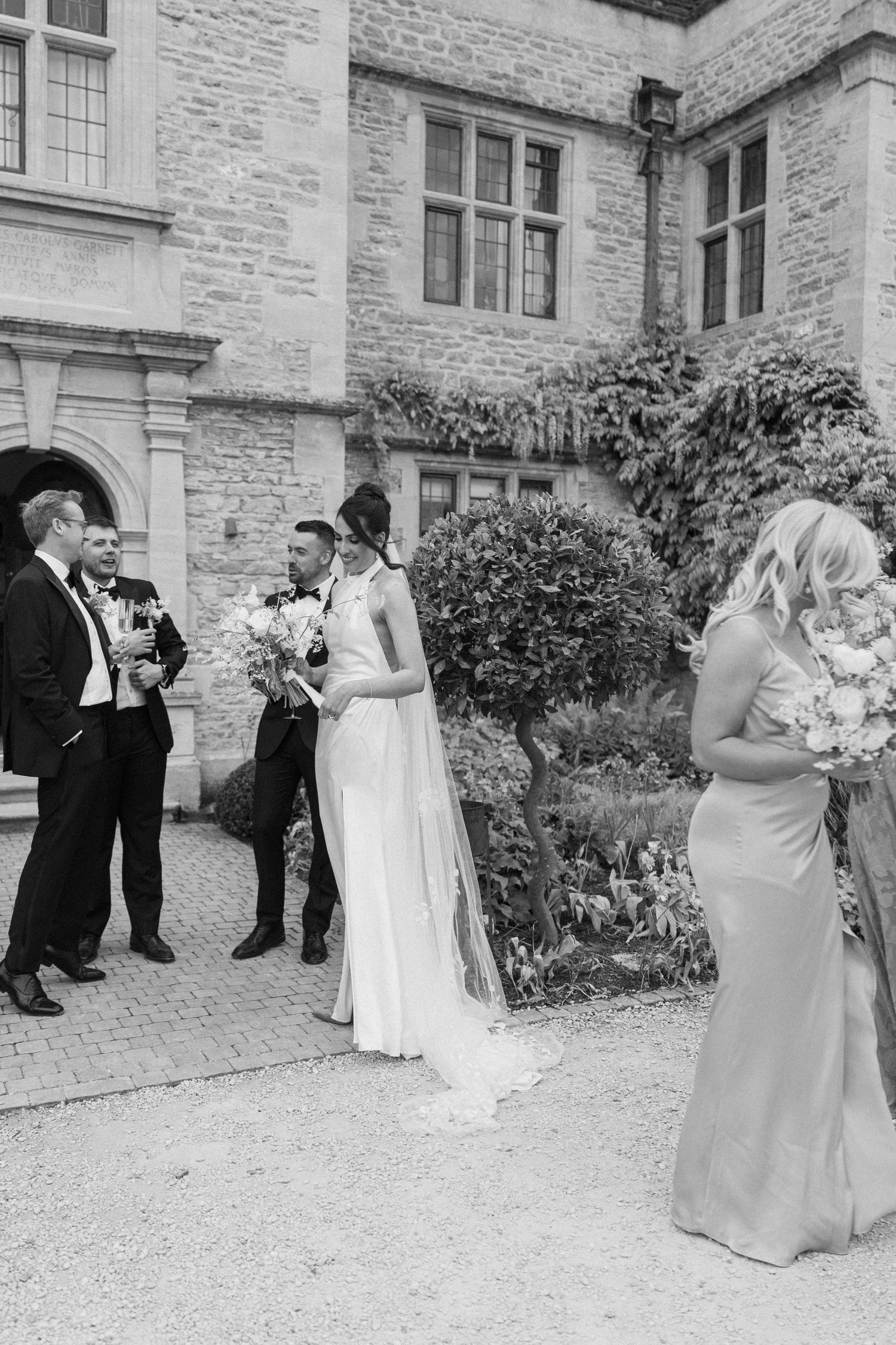 Black and white photo of wedding party gathered outside stone manor, bride in white gown with veil and bridesmaids.