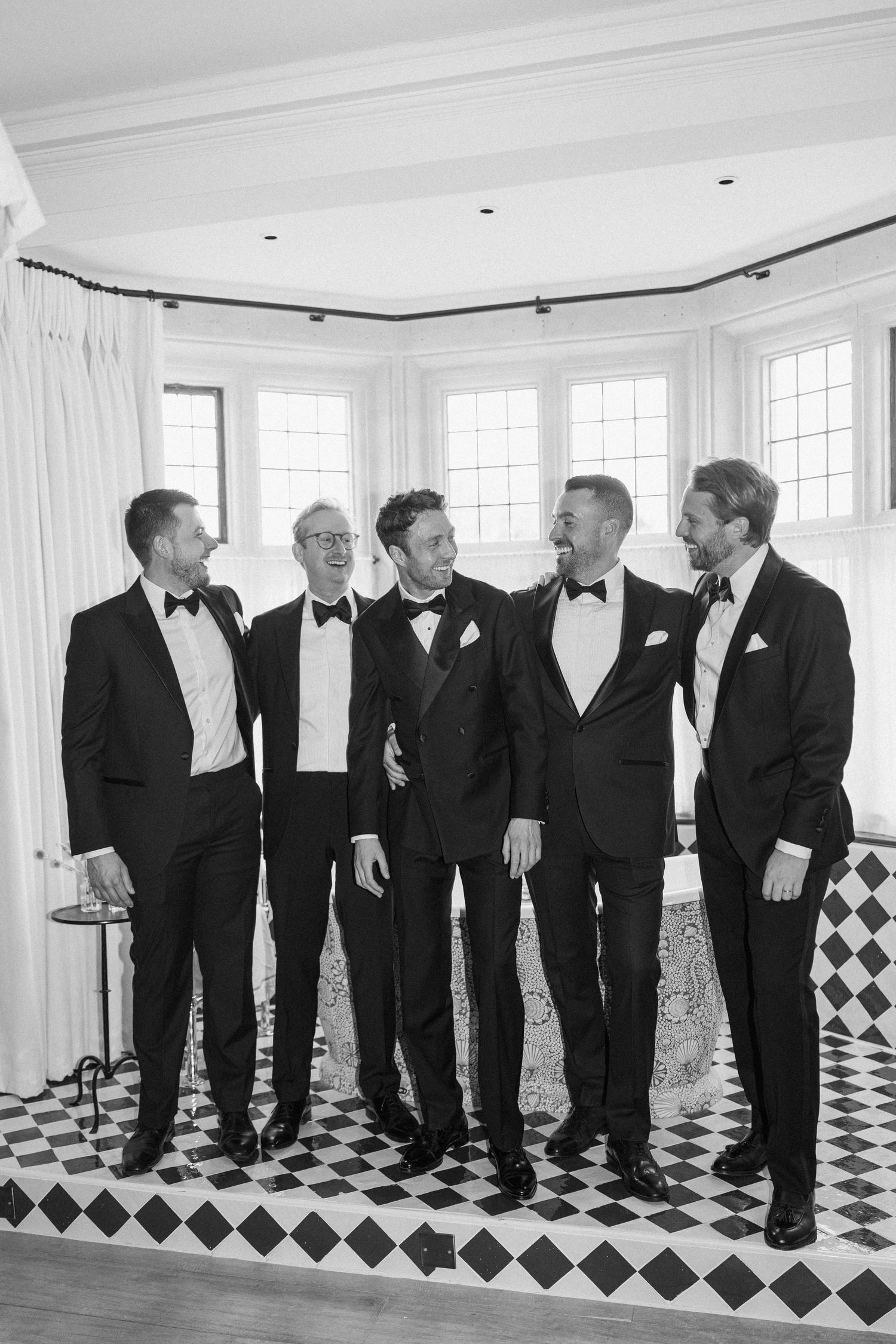 Black and white photo of five groomsmen in tuxedos laughing together in a bright bay window room.