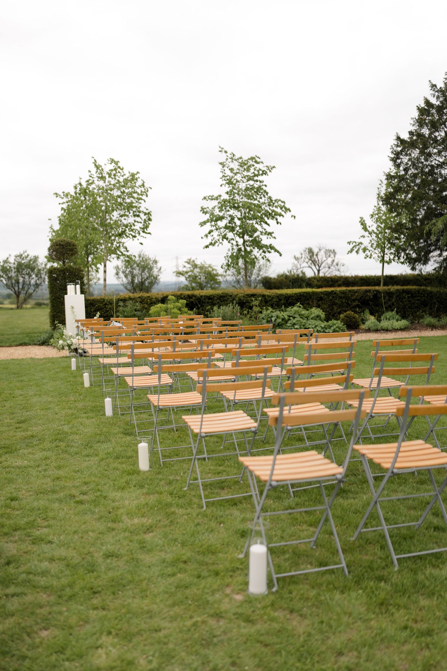 Rows of wooden folding chairs set up on green lawn for an outdoor wedding ceremony with candles and trees.