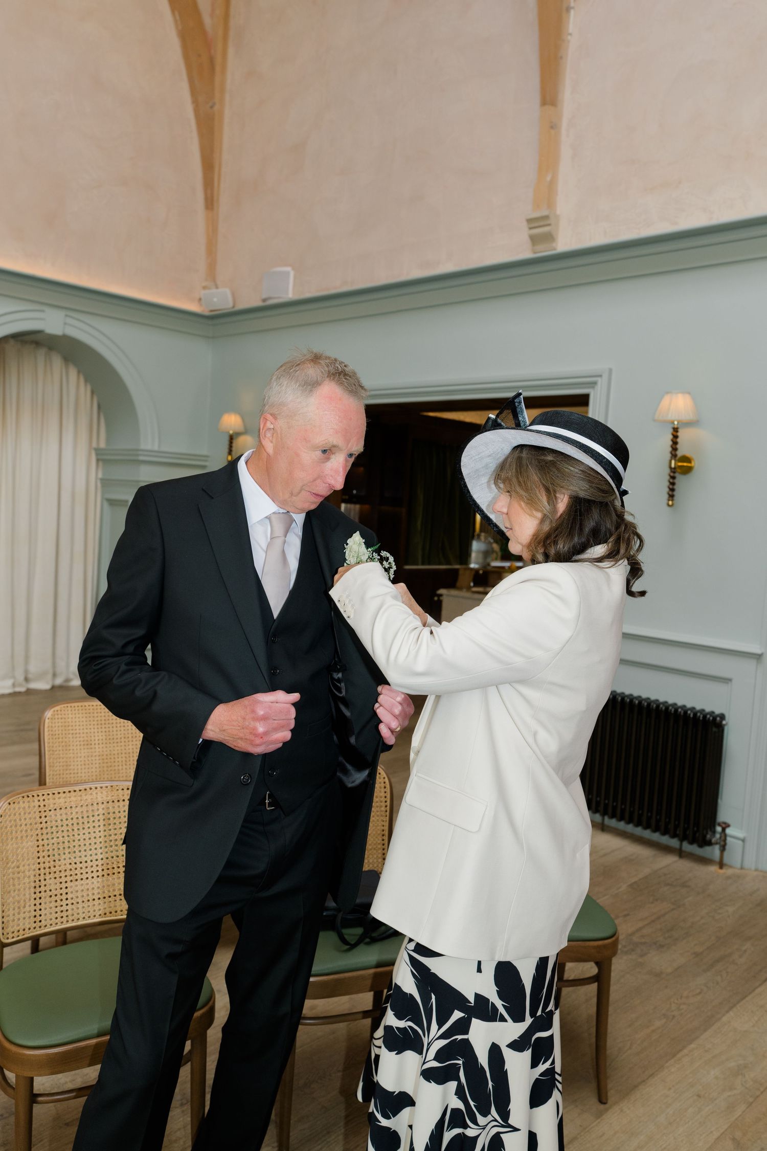 An older gentleman has his boutonniere adjusted by a woman in a white jacket and black hat at a wedding.
