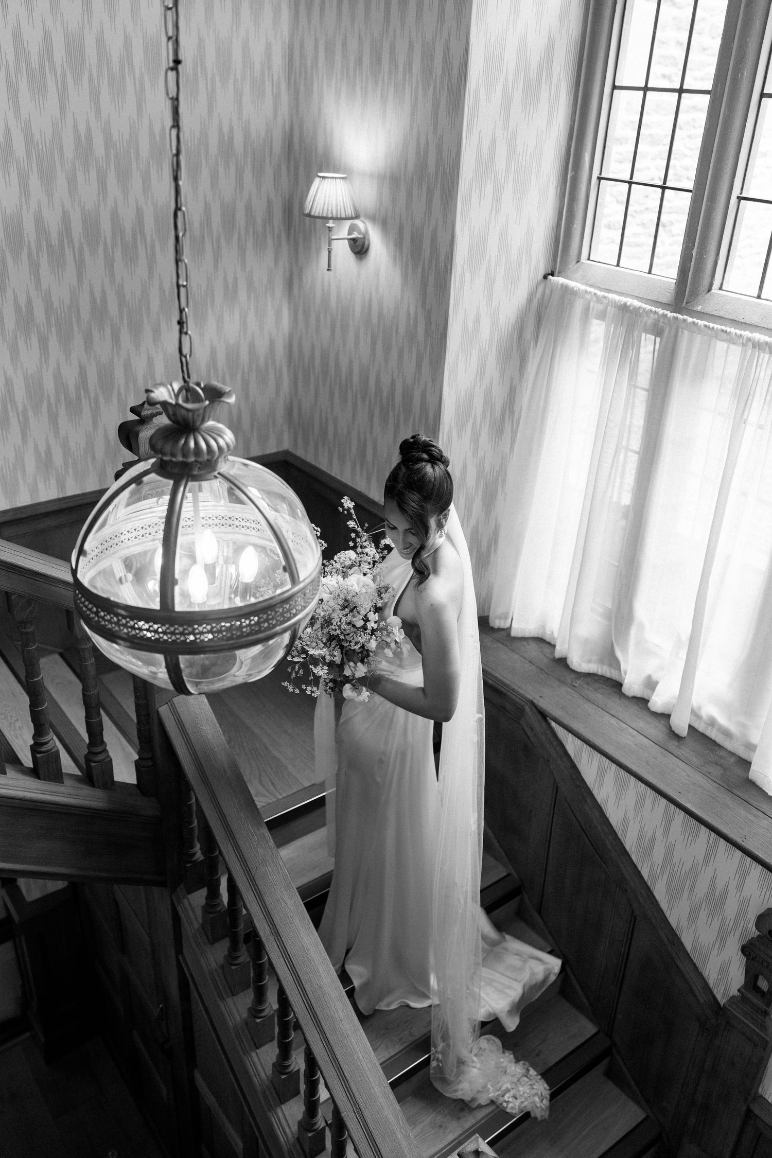 Bride in white gown holding bouquet descends elegant staircase in black and white wedding photo.
