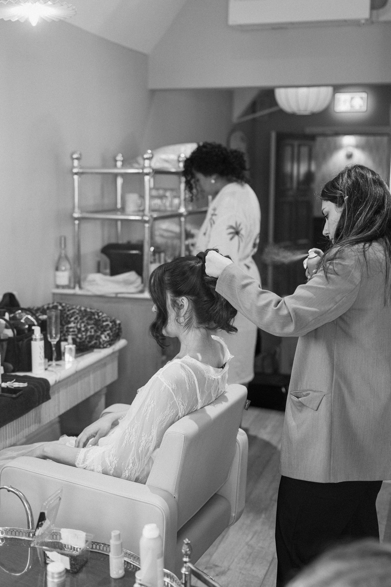 Black and white photo of a bride having her hair styled by two hairdressers in a busy wedding preparation salon.