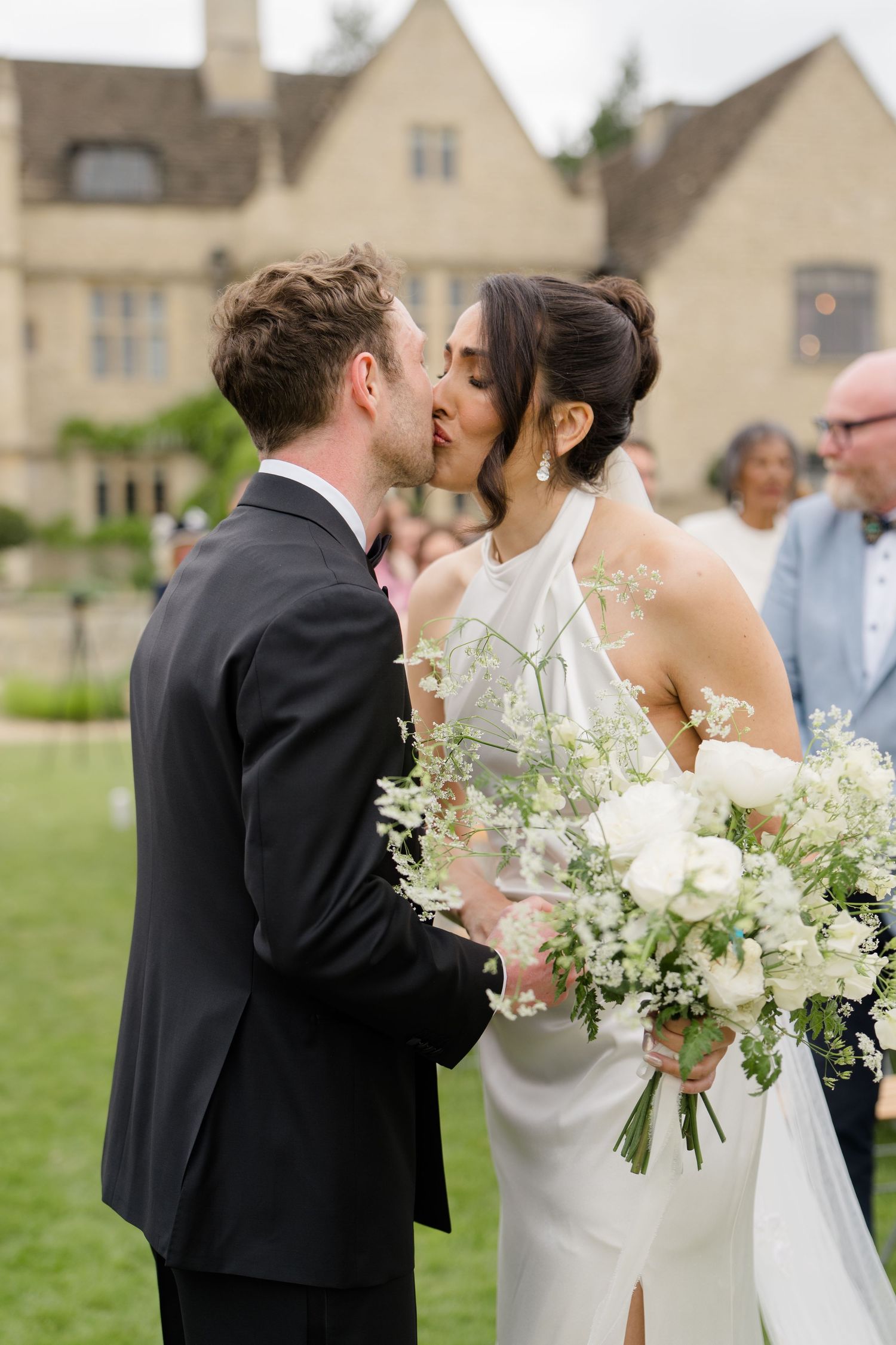 Newlyweds share first kiss as bride holds white floral bouquet outside elegant stone manor house.