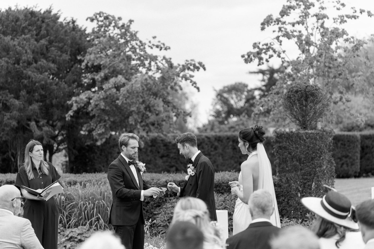 Groom exchanges vows with bride during elegant outdoor garden wedding ceremony before lush hedgerows.