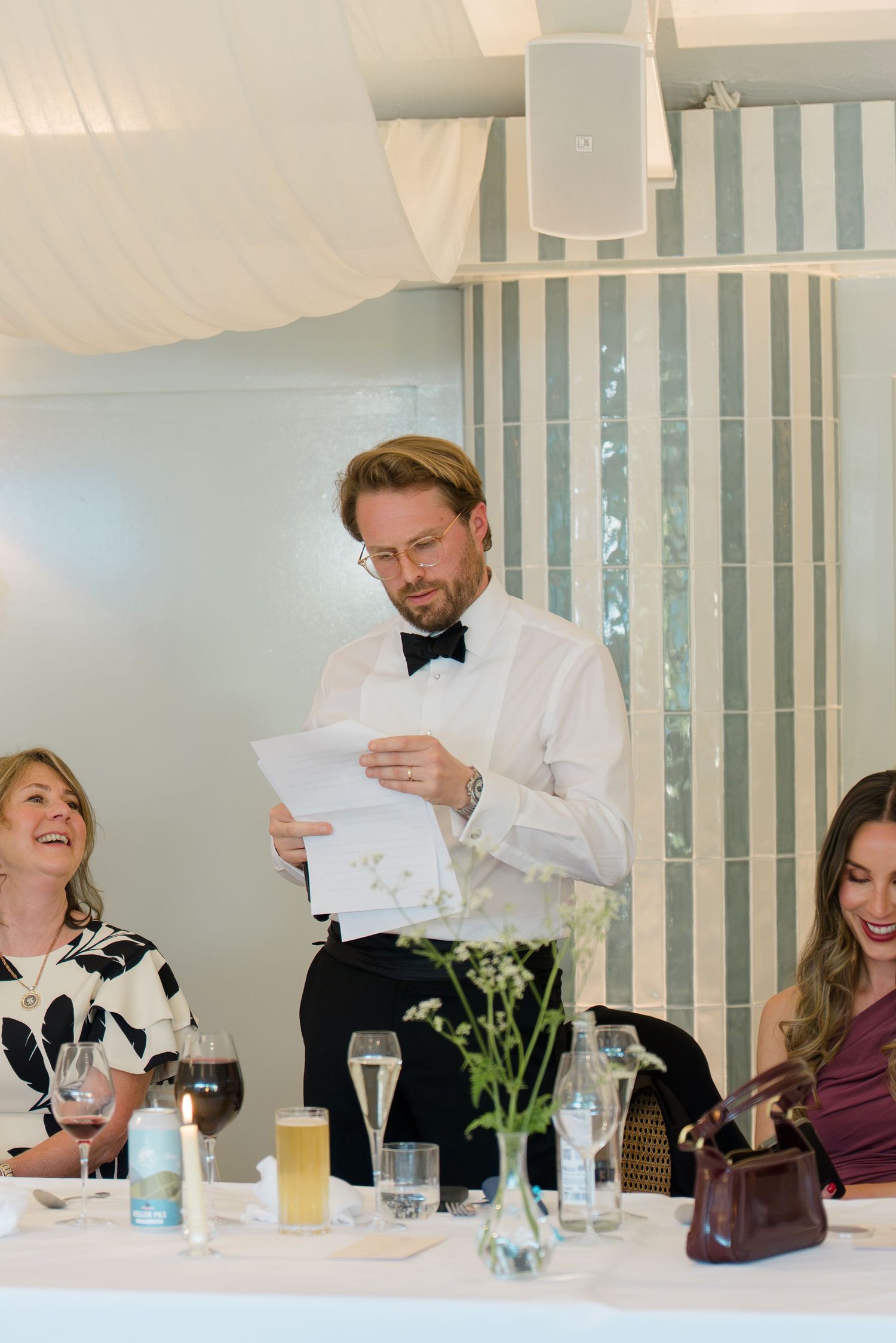 Best man in bow tie delivers a heartfelt wedding speech to laughing guests at the reception dinner.