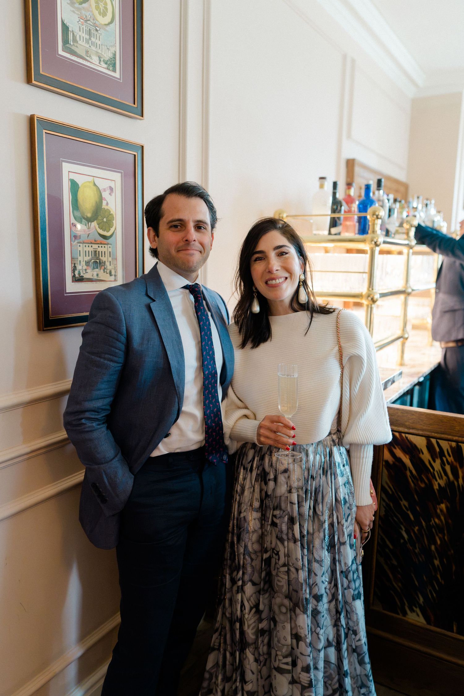 Elegant wedding guests pose together near a bar cart, man in blue suit and woman in floral skirt.