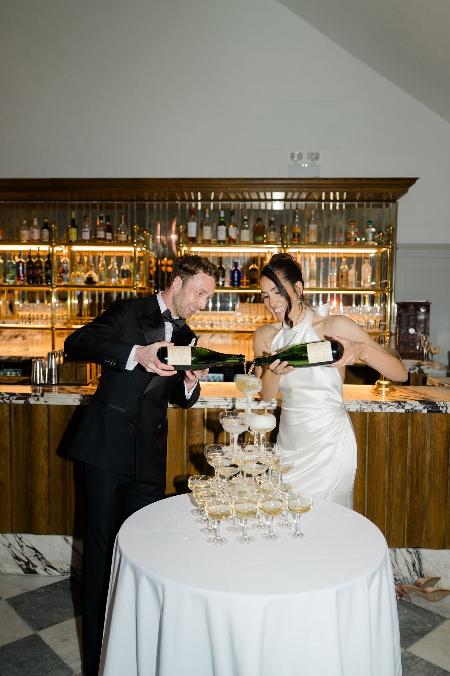 Bride and groom laugh while pouring champagne over a towering champagne tower at the bar.