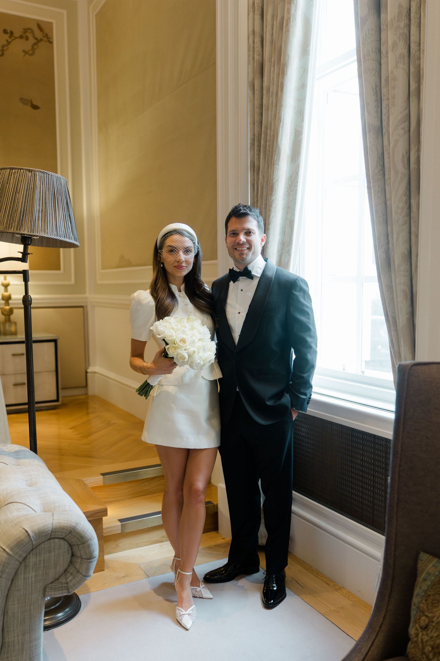 Elegant bride in white mini dress holding bouquet poses with groom in black tuxedo indoors.