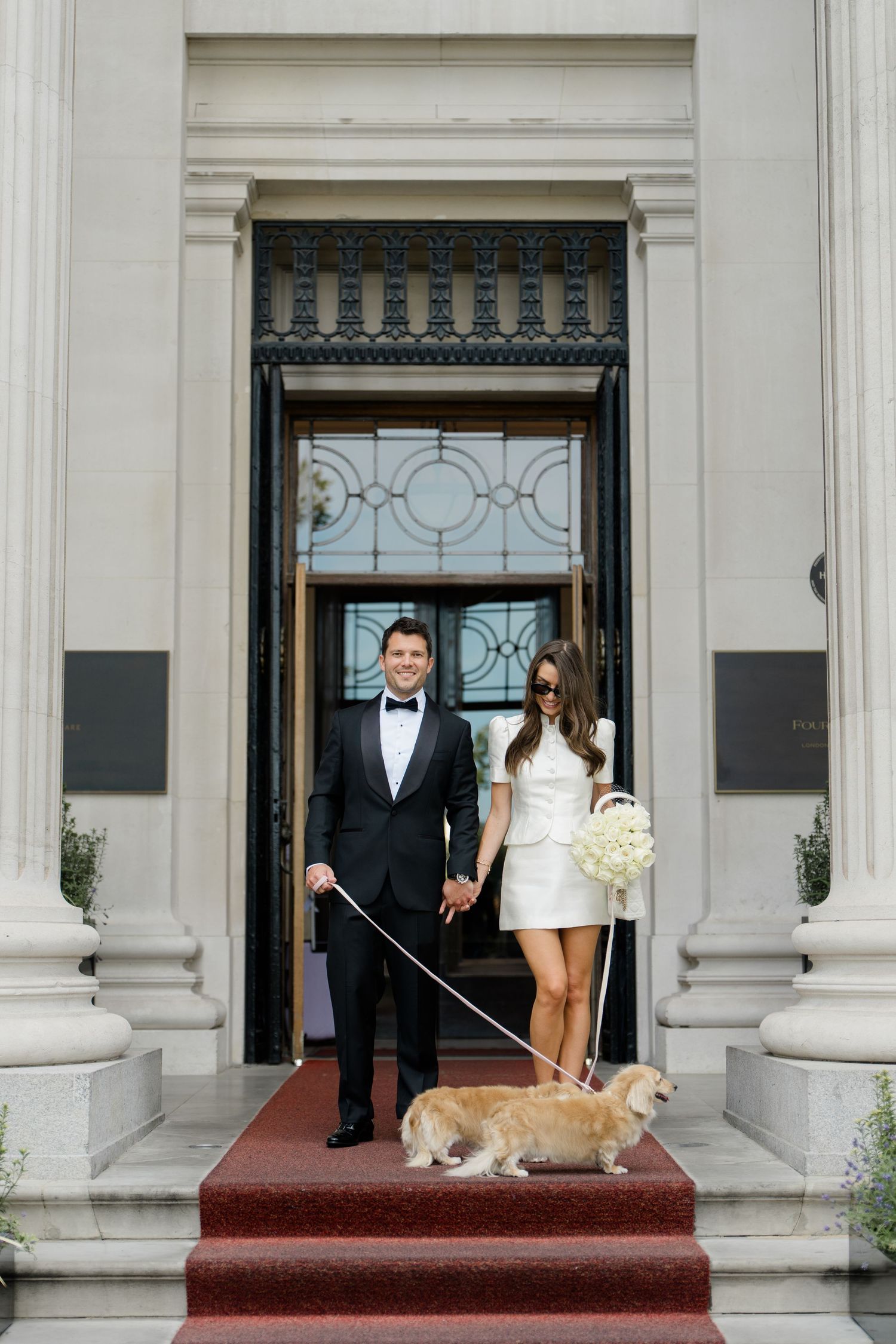 Bride and groom hold hands with their corgi on red carpet steps outside a grand building.