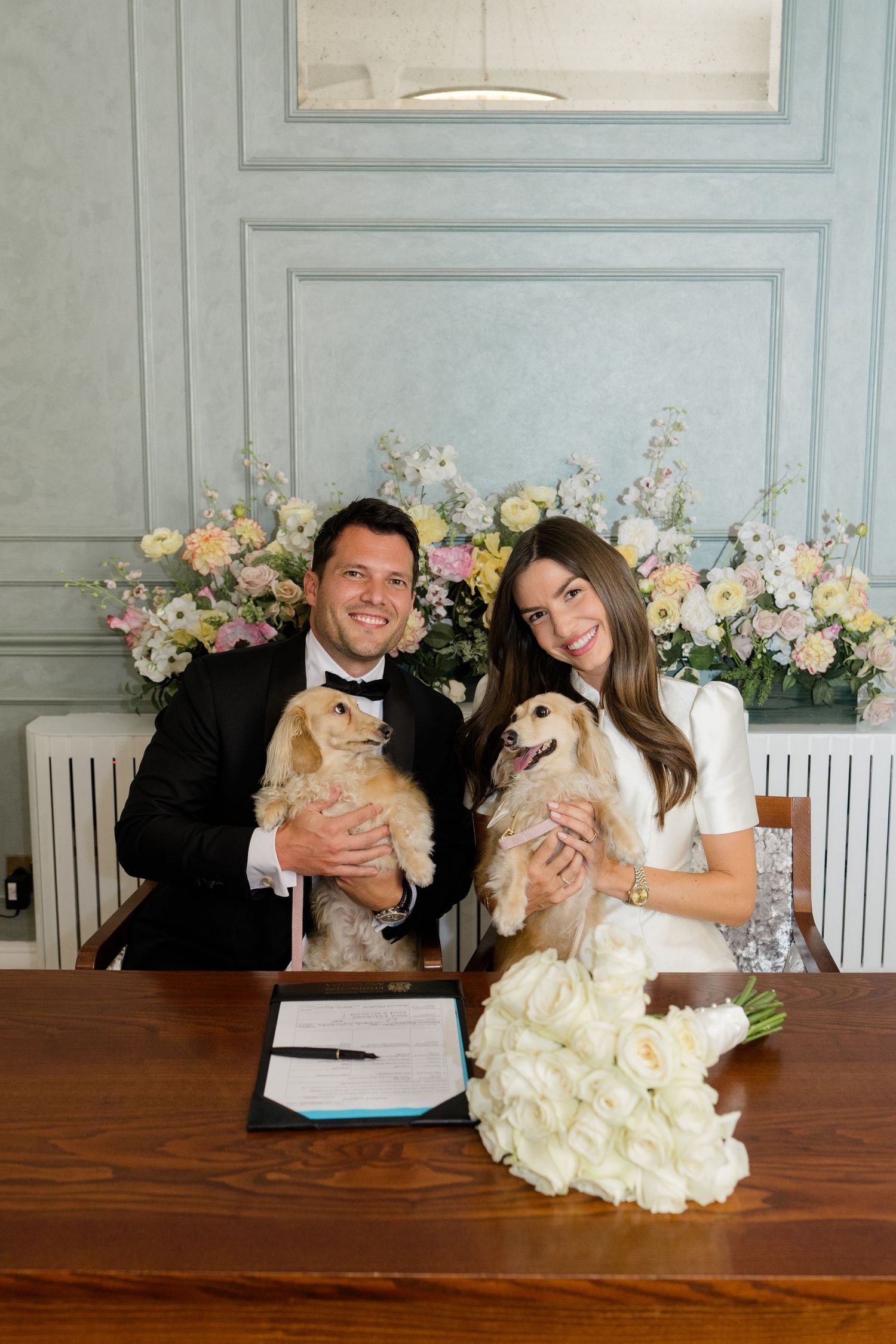 Smiling newlyweds holding cocker spaniel dogs at signing table with white tulip bouquet and flowers.