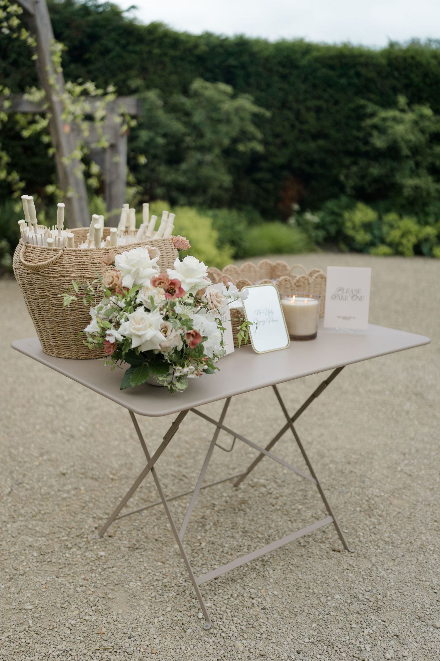 Elegant wedding welcome table with wicker basket, white and pink floral arrangement, candle, and signage on gravel path.