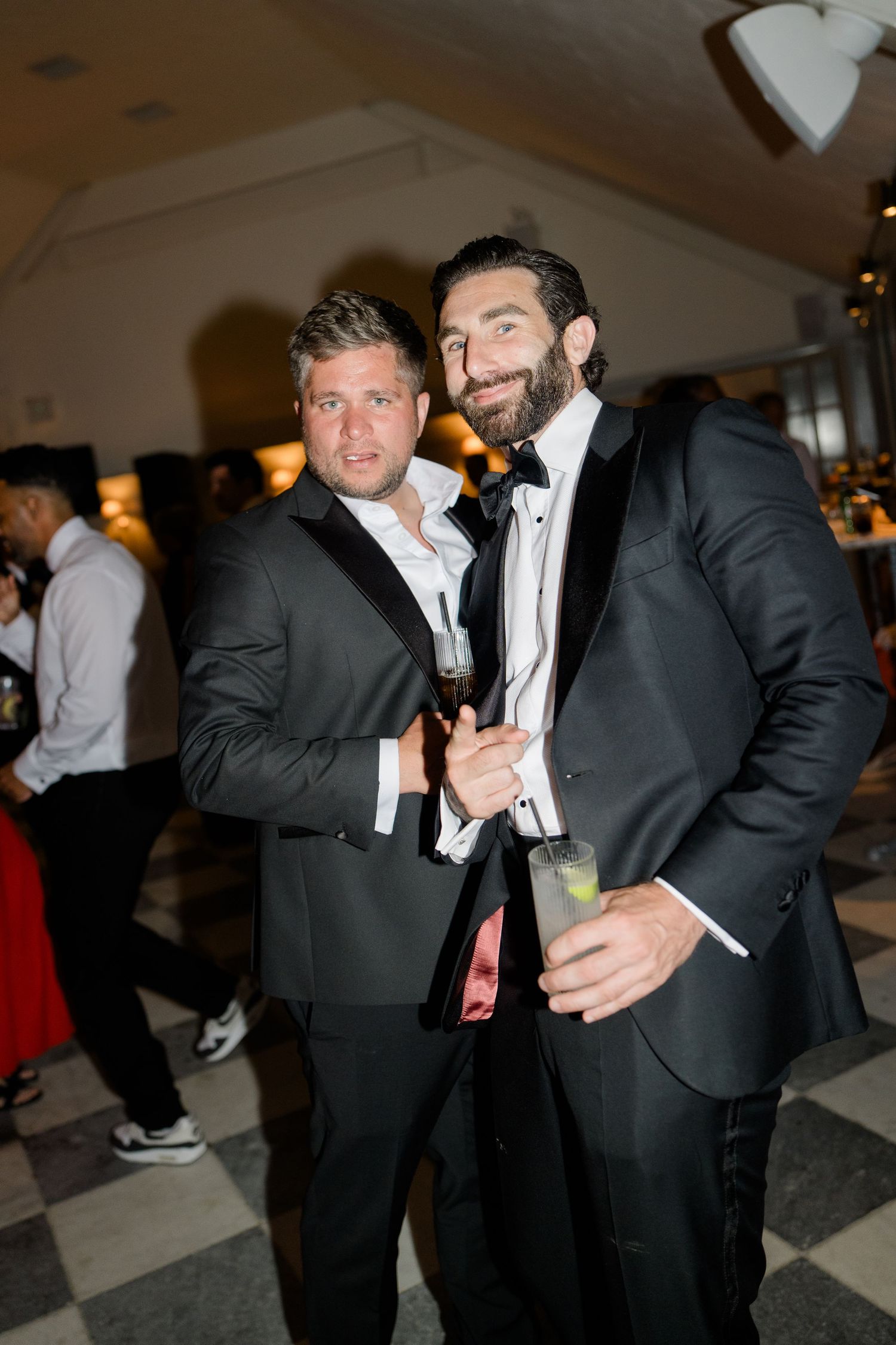 Two men in black tuxedos posing together holding drinks at a formal wedding reception party.