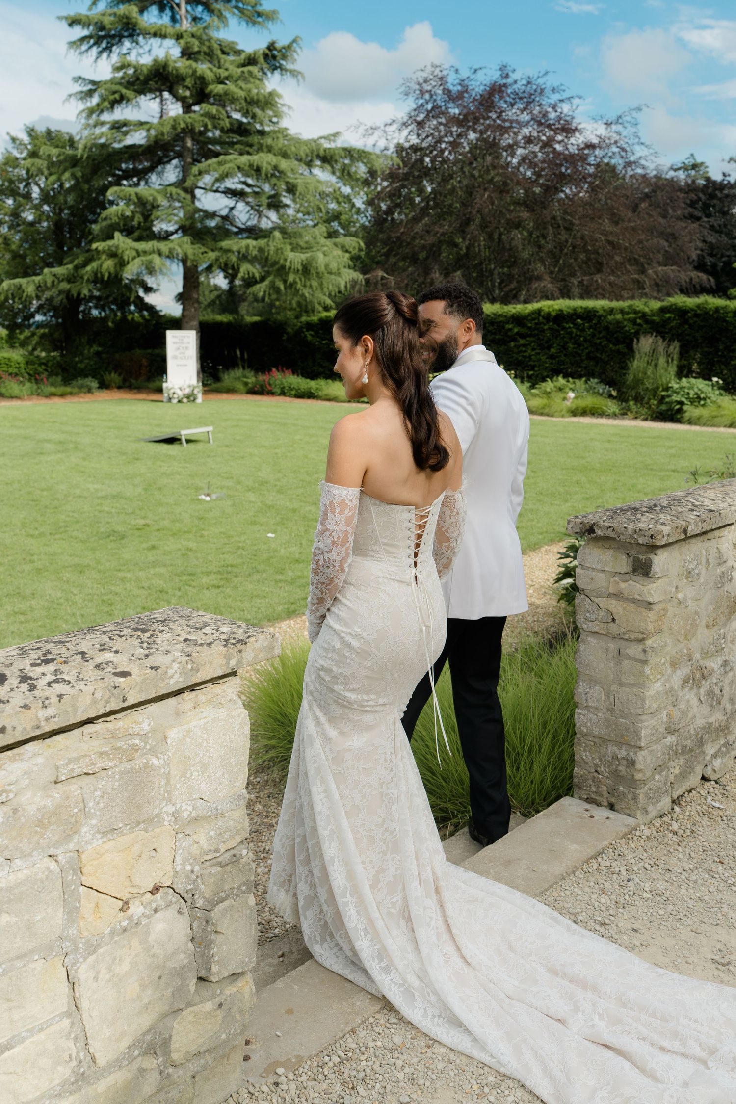 Bride in backless lace gown with train stands with groom in white jacket in elegant garden setting.