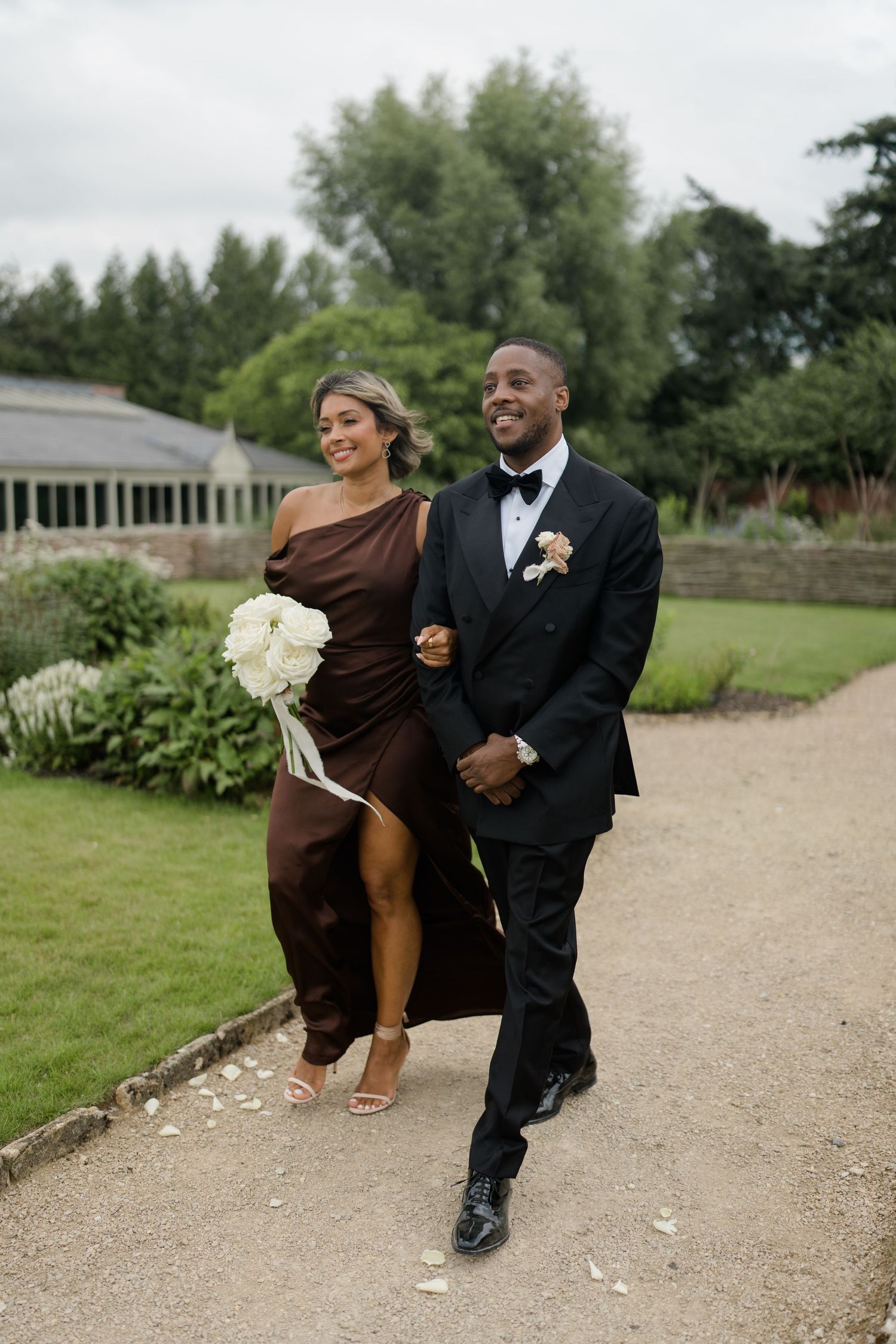 Bridesmaid in brown one-shoulder gown and groomsman in black tuxedo walking with white bouquet.