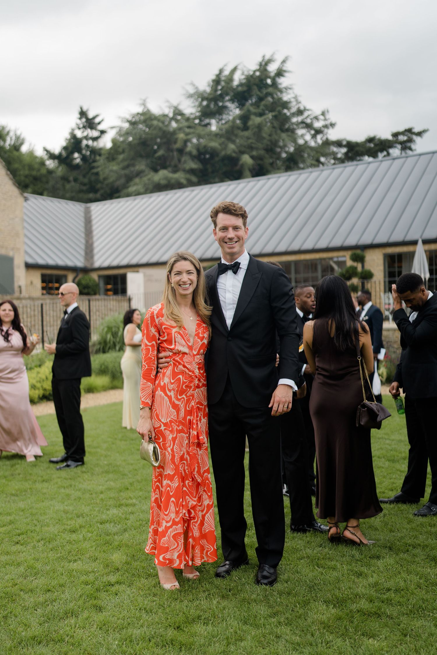 Couple posing at wedding reception, woman in orange print dress, man in black tuxedo.