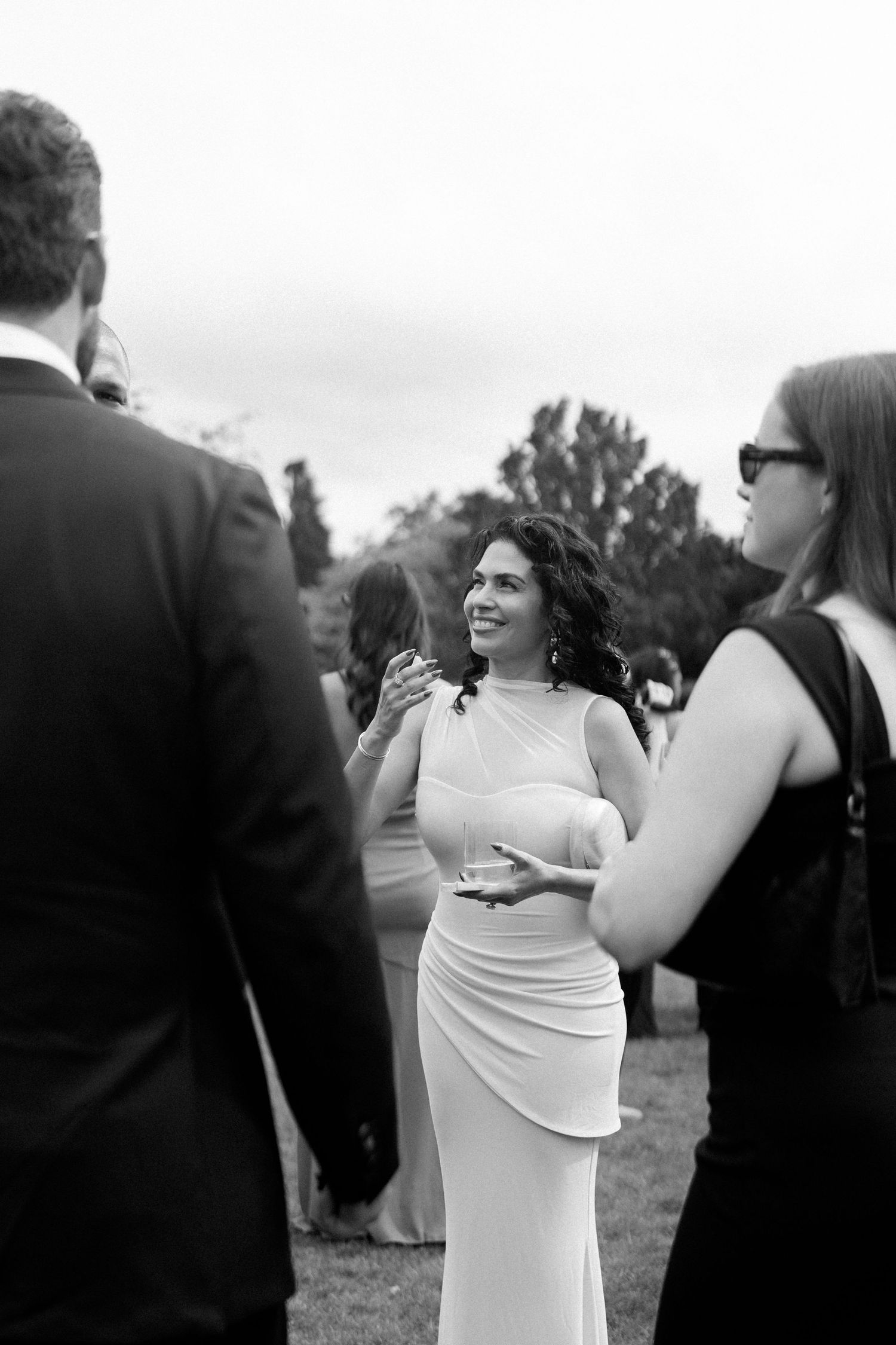 Black and white photo of a smiling woman in a white gown conversing with guests at an outdoor wedding.