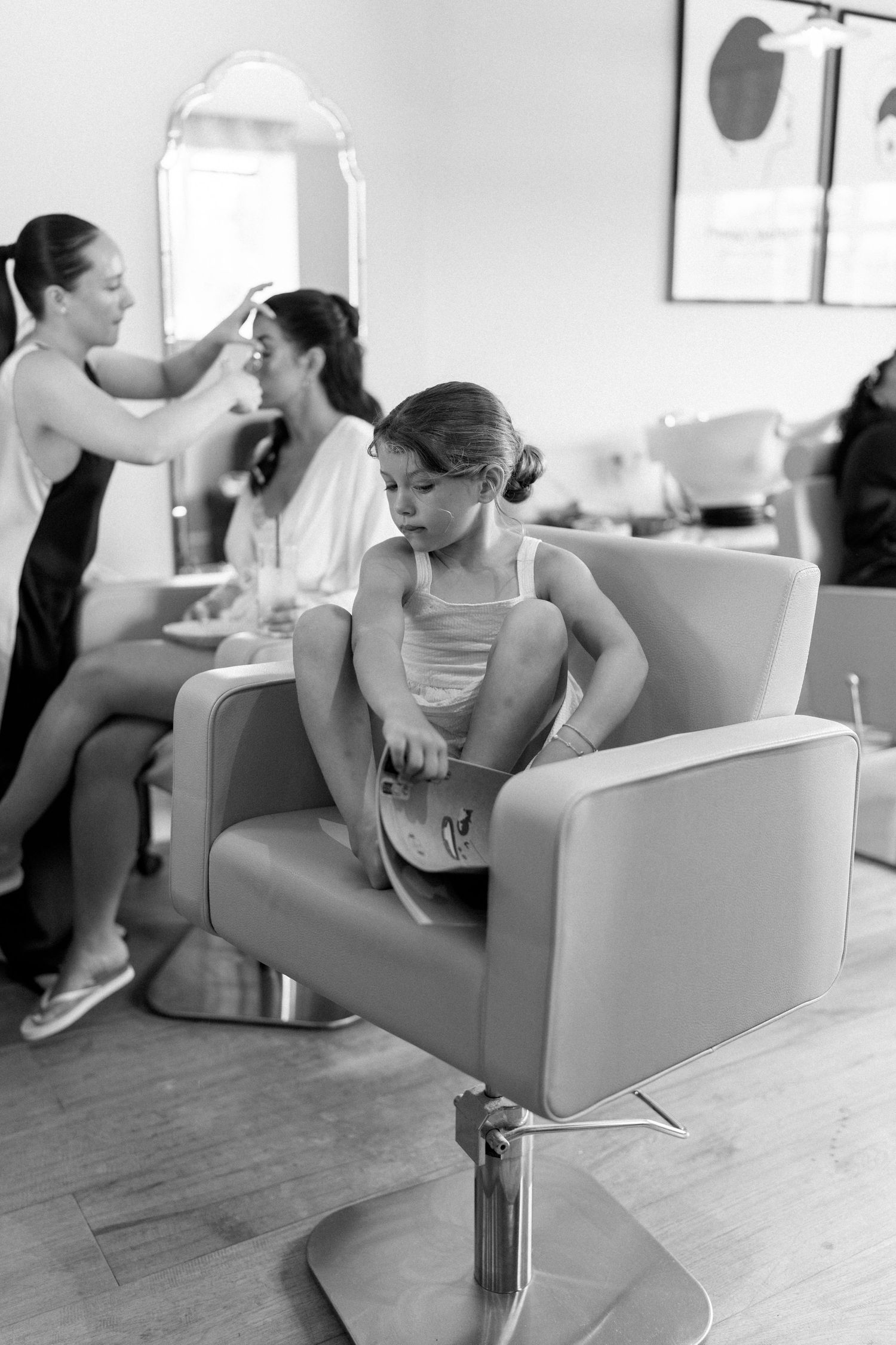 Young flower girl sitting alone in a salon chair during bridal party preparations, black and white photo.