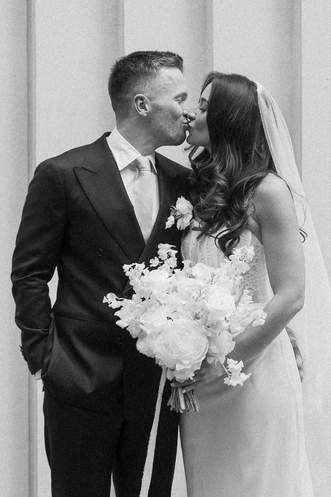 Black and white photo of a bride and groom sharing a kiss, bride holding a white floral bouquet.