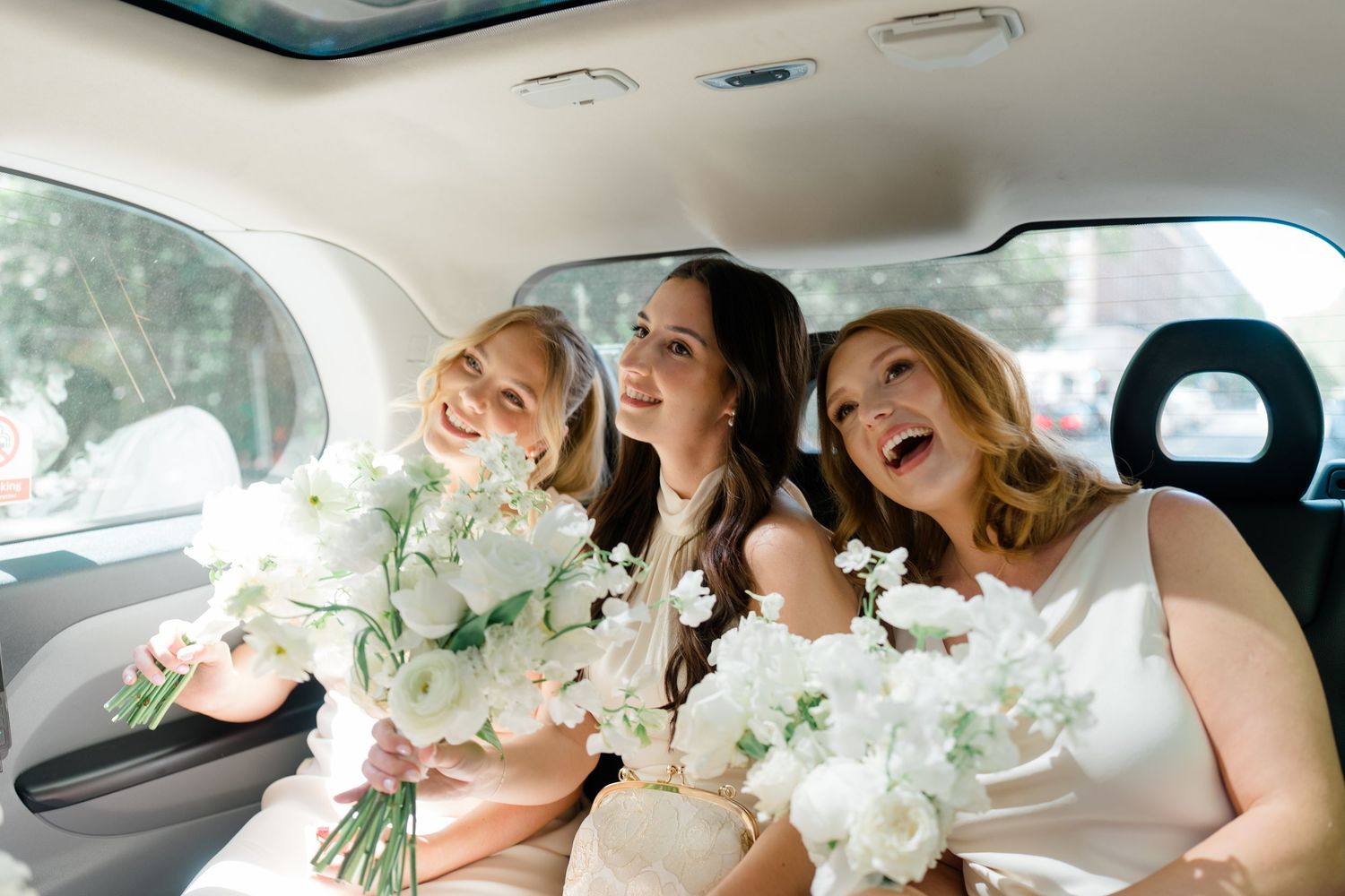 Three joyful bridesmaids holding white floral bouquets while sitting together in a wedding car.
